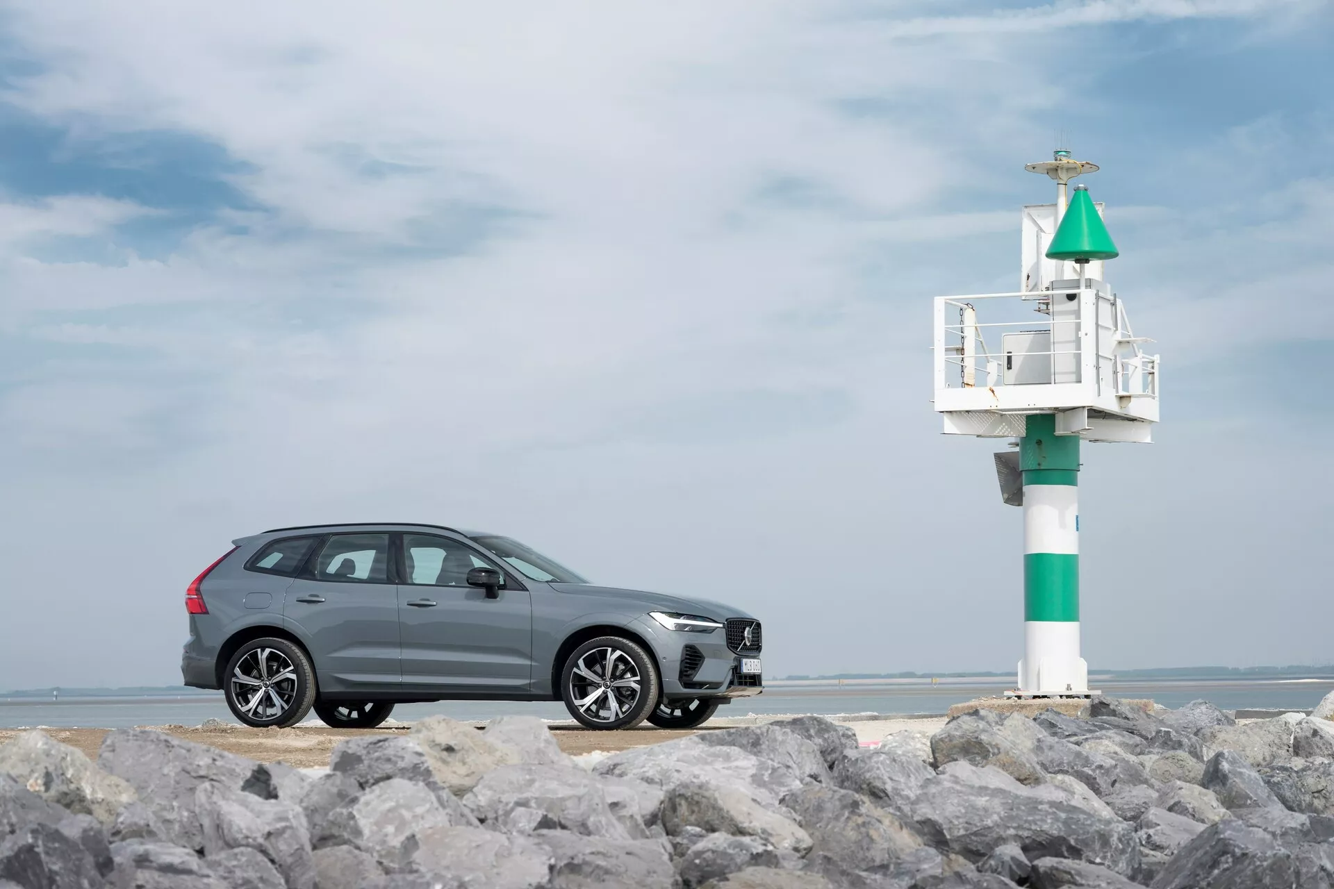 Vue de profil d’un SUV Volvo XC60 2021 gris (2ème génération, lifting) sur un sol rocailleux avec une balise maritime en arrière-plan. Le design élégant comprend des garnitures de fenêtre chromées, des jantes en alliage à plusieurs branches et un signal de virage intégré au rétroviseur latéral, capturé sous un ciel côtier partiellement nuageux.