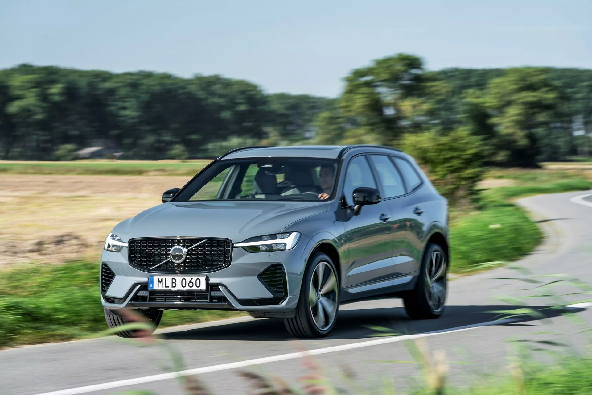 Prise de jour dynamique d'un Volvo XC60 2021 (deuxième génération-restylage) SUV en mouvement capturée sous un angle frontal de trois quarts. L'image montre la calandre avant avec un emblème Volvo proéminent, des phares à LED angulaires, des lignes aérodynamiques et des jantes en alliage modernes à rayons multiples. Les feux de jour sont allumés, indiquant que la voiture est en mouvement. La photo met en avant le design élégant et la présence sur route de ce véhicule utilitaire familial ou de luxe moderne.