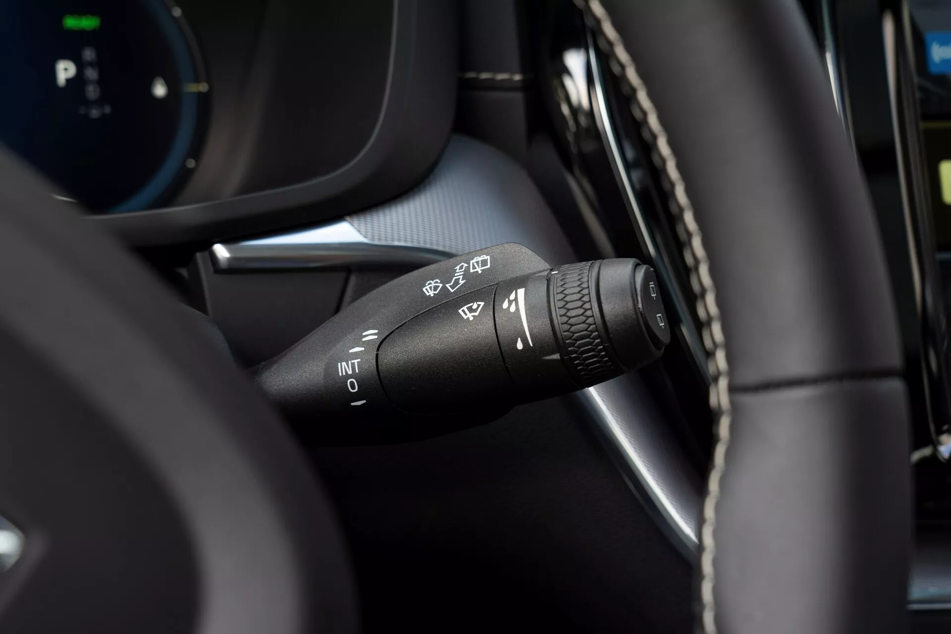 Gros plan sur le levier de commande des essuie-glaces de la Volvo XC60 2021 (2e génération restylée) depuis la perspective du conducteur. Axé sur les divers symboles de réglage des essuie-glaces : détection automatique de la pluie, intermittent, basse vitesse et haute vitesse. La pointe du levier comporte une bague rotative pour ajuster l'intervalle des essuie-glaces intermittents. Intérieur moderne avec un mélange de matériau ressemblant à du cuir et des composants en plastique noir, avec un tableau de bord numérique flou en arrière-plan.