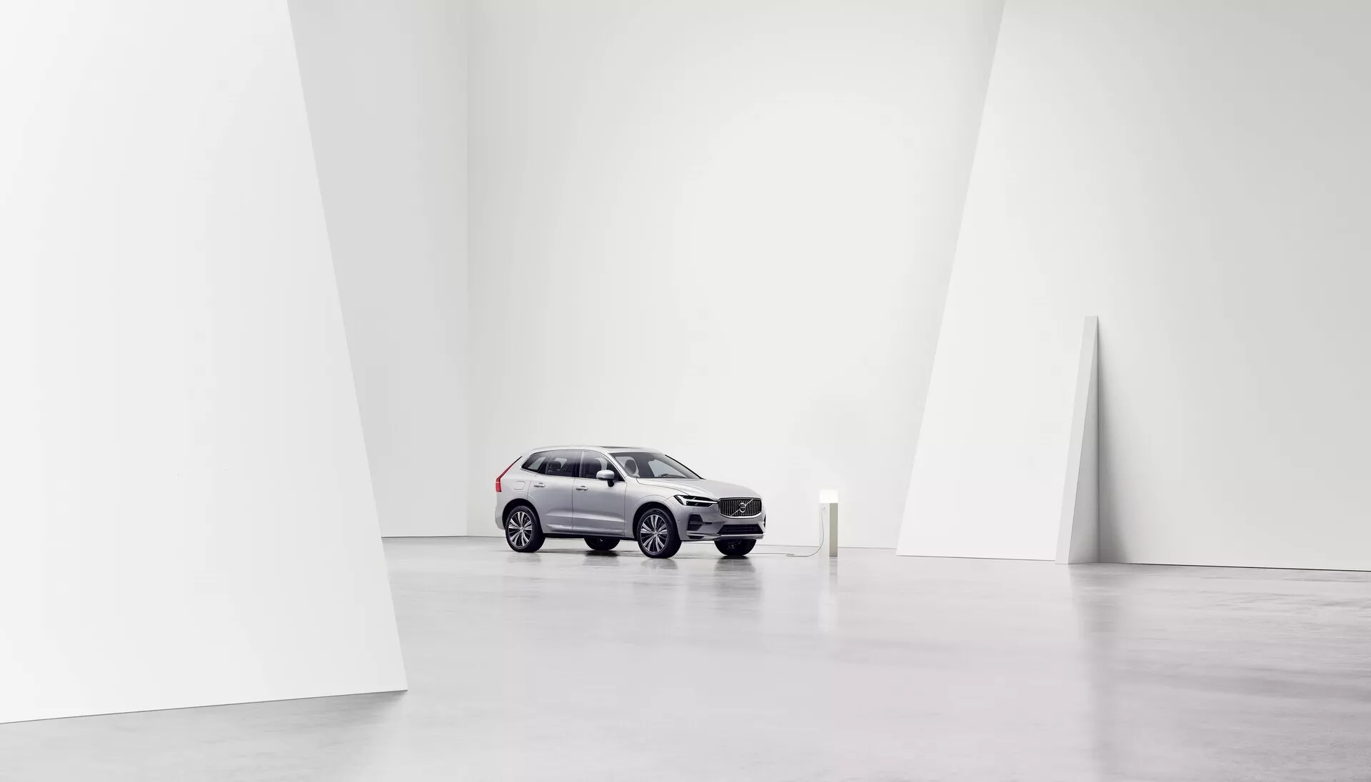 Image d'une Volvo XC60 2021 (2e génération, facelift) positionnée au centre d'un environnement de studio minimaliste, spacieux et bien éclairé, capturée sous une perspective de trois quarts avant. La couleur argent clair de la voiture contraste avec ses roues plus foncées et une partie de la calandre avant, montrant des éléments de design modernes de SUV comme des lignes courbes et angulaires. La vue met en évidence la calandre avant, les phares, le capot, le pare-brise et le côté conducteur, y compris les portes avant et arrière et les vitres latérales. Le fond blanc immaculé met en valeur la voiture, en focalisant l'attention sur son design et ses caractéristiques, idéal pour des fins promotionnelles ou artistiques.