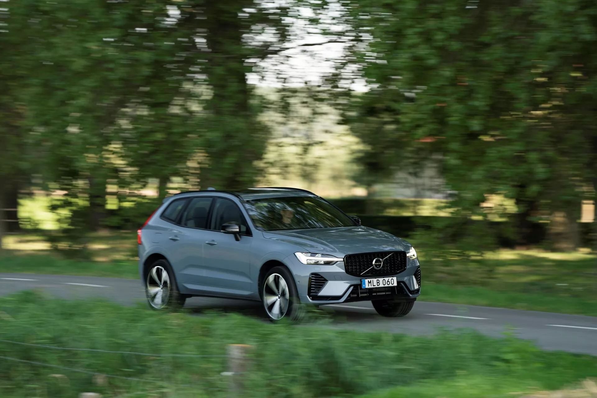Break moderne Volvo XC60 2021 de couleur grise en mouvement, capturé sous un angle latéral sur une route de banlieue ou de campagne avec de la verdure de chaque côté. Le quart de panneau avant droit, le côté et une partie arrière sont visibles. Mouvement dynamique accentué par la technique du panoramique, arrière-plan et roues flous, détails de la voiture nets incluant la calandre distinctive, phares élégants, lignes aérodynamiques et poignées de porte affleurantes. Design visible à distance moyenne, plaque d'immatriculation floue, montrant le design et le mouvement de la Volvo dans un cadre naturel.