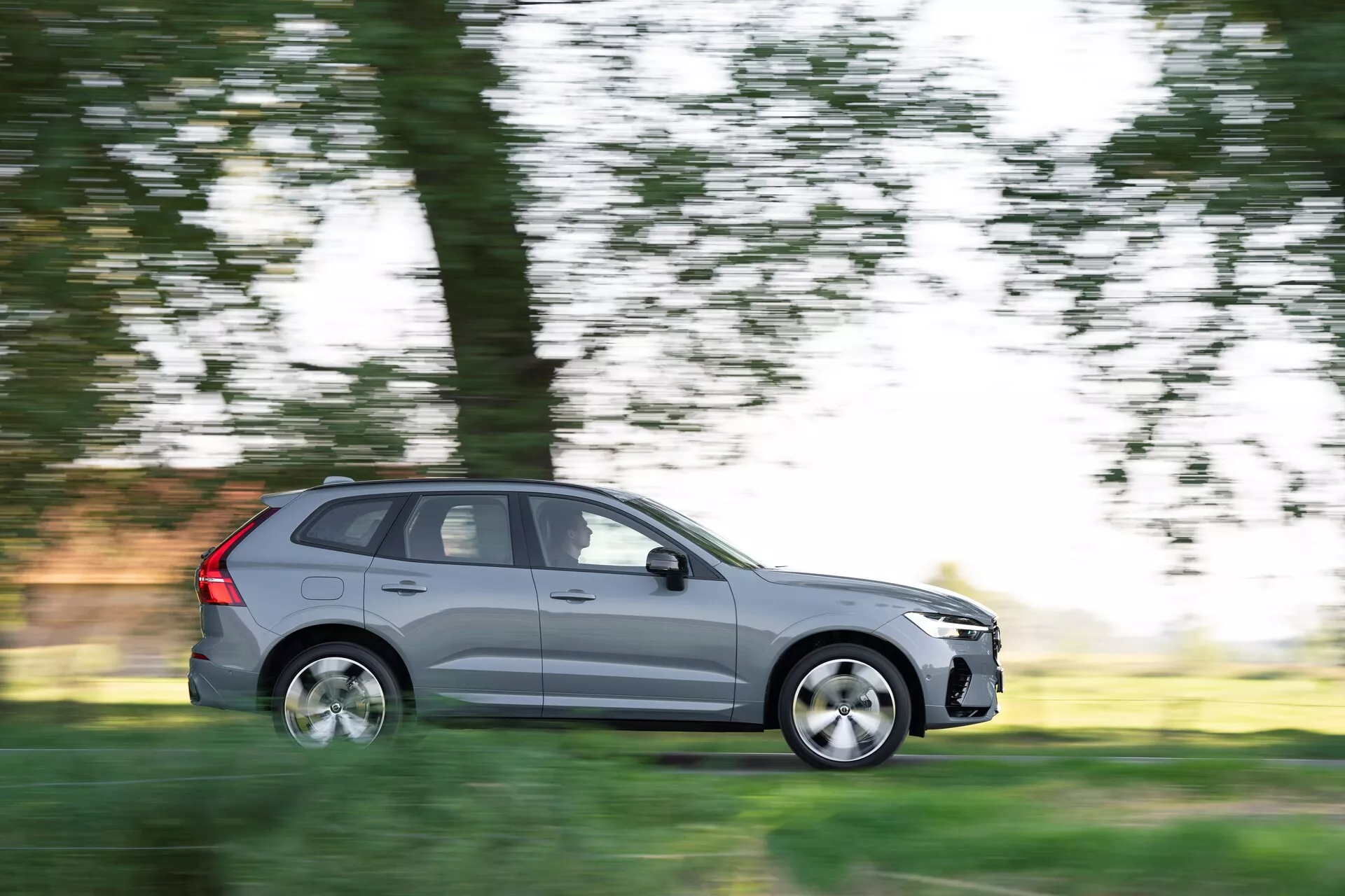 L'image présente un SUV Volvo XC60 2021 (2ème génération, facelift) en mouvement, capturé avec une technique de panoramique qui floute l'arrière-plan tout en gardant la voiture nette. Vue du côté conducteur en profil latéral complet, le véhicule gris métallisé présente cinq portes, des roues en alliage et des pneus standard pour la route. La scène suggère une conduite diurne à travers un environnement naturel, avec des arbres et de l'herbe flous en arrière-plan, mettant en valeur le mouvement de la voiture.