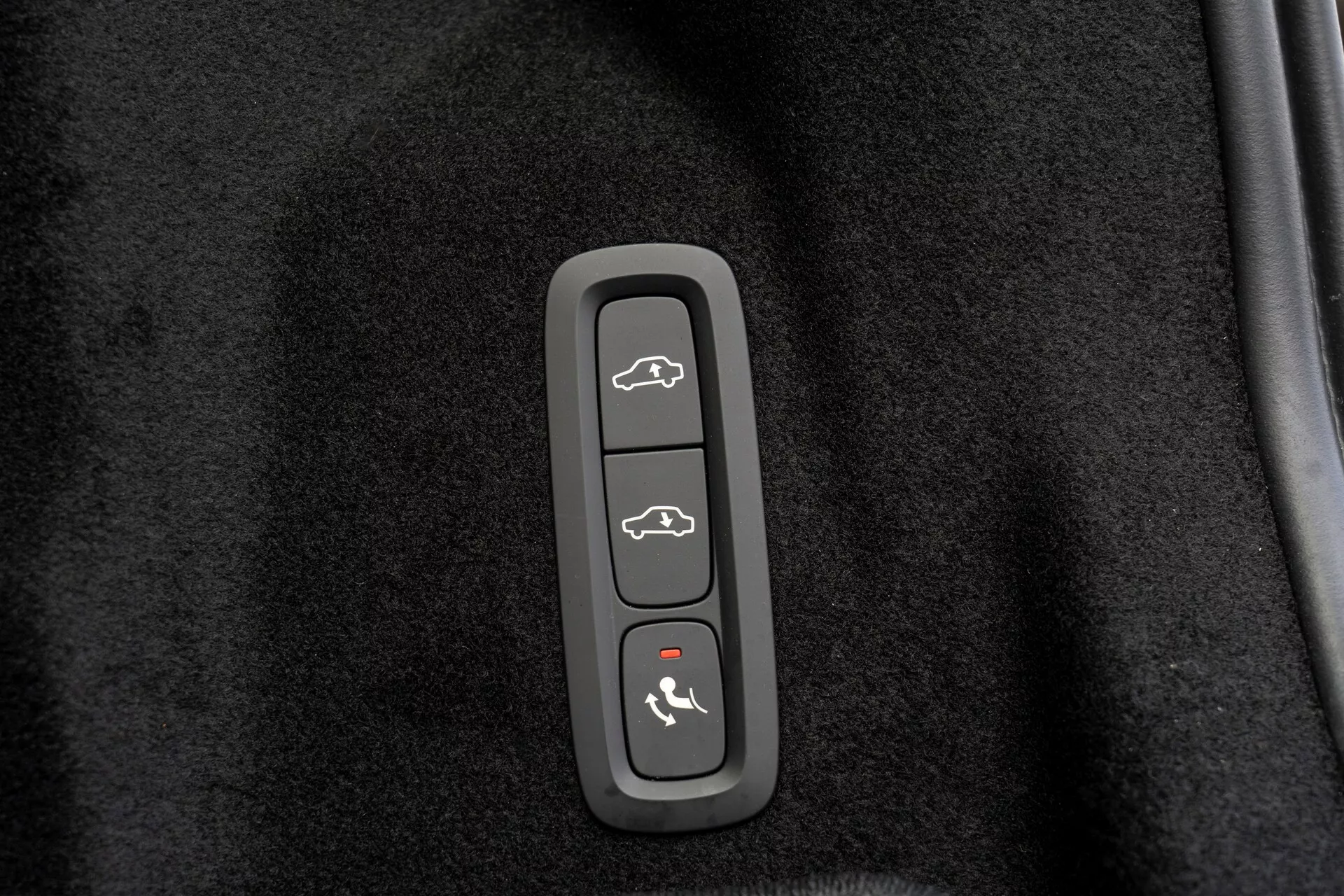 Gros plan d'un module de commande dans une Volvo XC60 2021 (2e génération, restylée), intégré dans une garniture en plastique contre un fond noir ressemblant à un tapis. Le module comporte quatre boutons : en haut - véhicule avec coffre ouvert, deuxième - silhouette d'une voiture avec toutes les portes ouvertes, troisième - voiture avec une flèche pointant vers le capot, en bas - voiture avec une ligne courbe au-dessus, probablement pour l'ouverture du coffre, le verrouillage/déverrouillage des portes, l'accès au capot/mode valet et l'activation du système de sécurité ou l'alarme de panique.