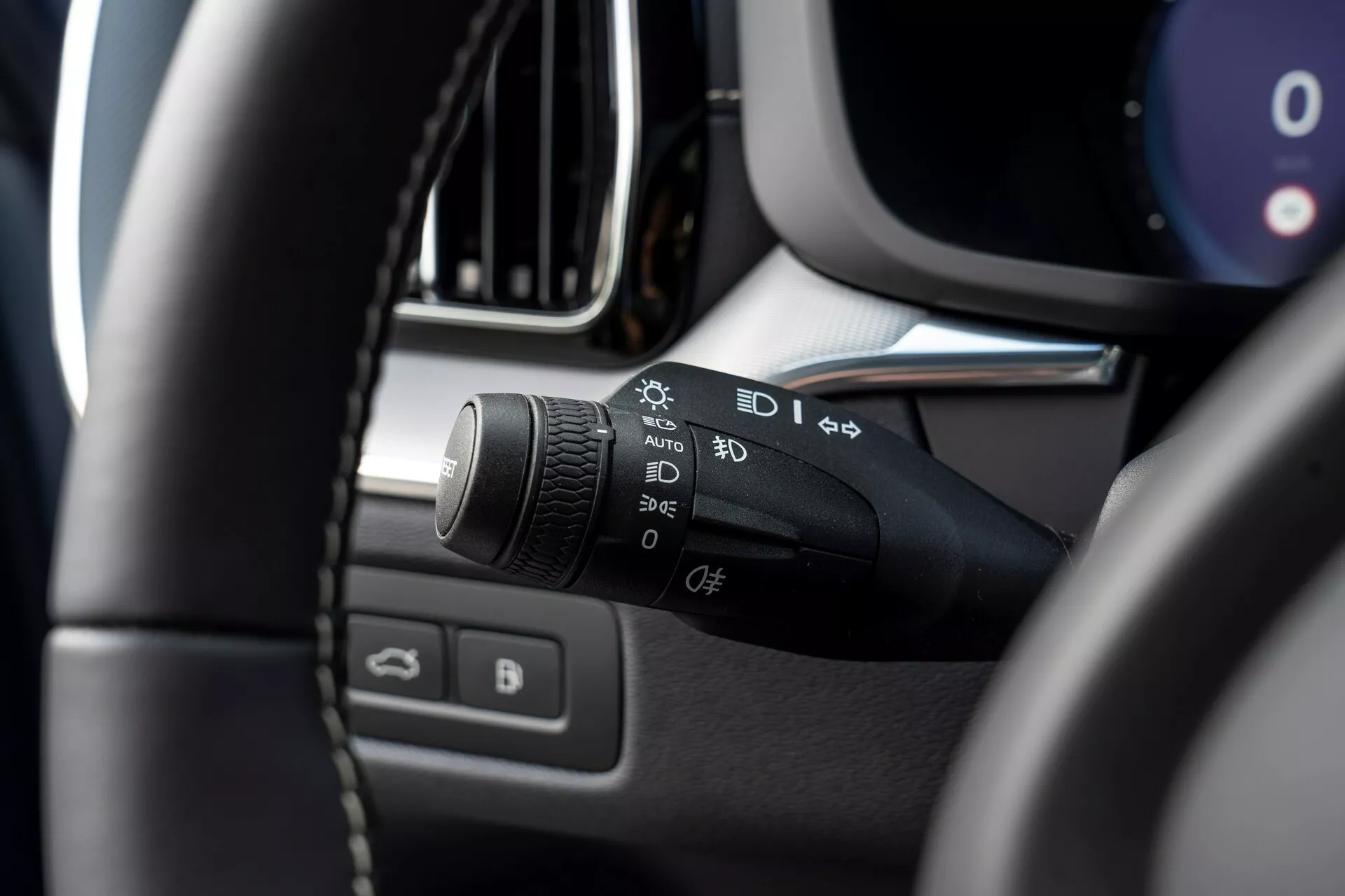 Gros plan de l'intérieur d'une Volvo XC60 2021 (facelift de 2e génération), se concentrant sur la colonne de direction côté gauche. Le sujet principal est la manette de commande des phares et des clignotants avec des symboles pour les feux automatiques, feux de stationnement, phares, réglages des phares antibrouillard et une fonction personnalisable. L'image est prise du siège conducteur, légèrement vers le haut et la gauche, montrant la partie supérieure gauche du volant floue au premier plan et mettant en avant la manette de commande. D'autres détails flous incluent des parties du tableau de bord, la console centrale, la bordure de l'aération ou de l'écran d'infodivertissement, et la porte du conducteur avec des commandes de fenêtres et de rétroviseurs.