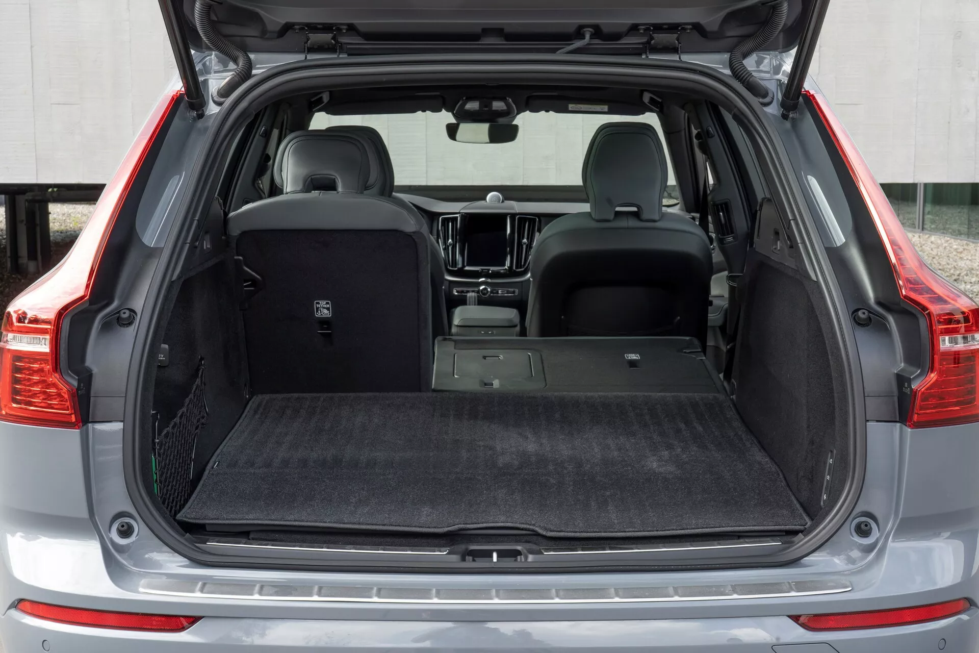 Vue arrière en gros plan d'une Volvo XC60 2021 deuxième génération restylée, avec le coffre ouvert pris sous un angle bas. L’image met en avant l’espace de chargement spacieux avec intrusion minimale des passages de roue, le couvre-bagages rétractable et le revêtement intérieur en moquette axé sur la réduction du bruit et la protection. Les sièges arrière partiellement rabattus montrent la polyvalence, tandis que la plaque protectrice sur le seuil de chargement, les capteurs de stationnement visibles sur le pare-chocs arrière, l'illumination LED du coffre et les points d’arrimage soulignent la fonctionnalité et l'attention aux détails.