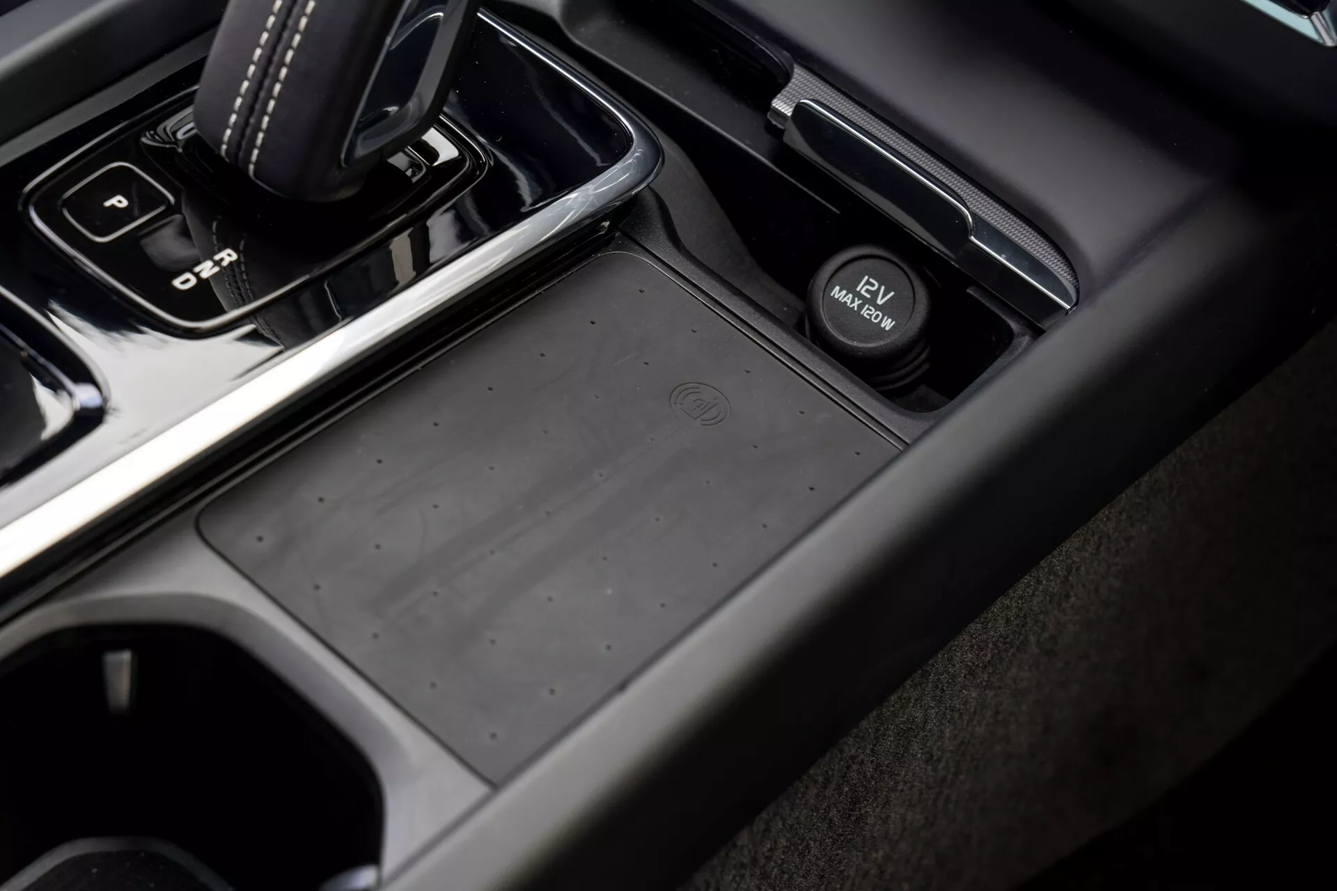 Vue rapprochée et angulaire de la zone de la console centrale d'une Volvo XC60 2021 (deuxième génération restylée) depuis le côté passager, avec un levier de vitesse automatique en position 'park', un compartiment de rangement antidérapant, un interrupteur de frein de parking électronique, des porte-gobelets et une combinaison de matériaux en plastique doux au toucher et finition métallique dans des tons gris foncé et noir.