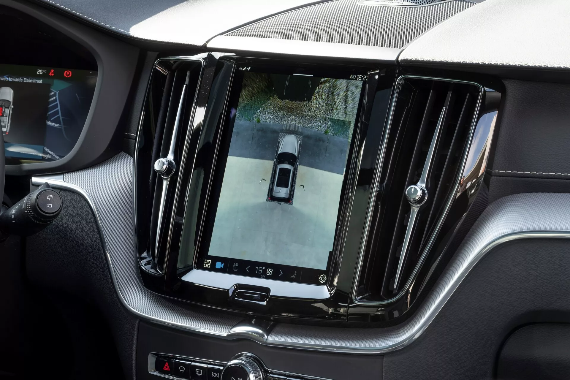 Vue intérieure d'une Volvo XC60 2021 (restylage de 2e génération) présentant un grand écran tactile d'infodivertissement vertical intégré dans la console centrale, affichant le flux de la caméra de recul avec une vue en plongée. Finition de luxe avec accents chromés et éléments de design épurés, commandes supplémentaires sous l'écran et vue partielle du volant, indiquant un intérieur premium et technologiquement avancé.
