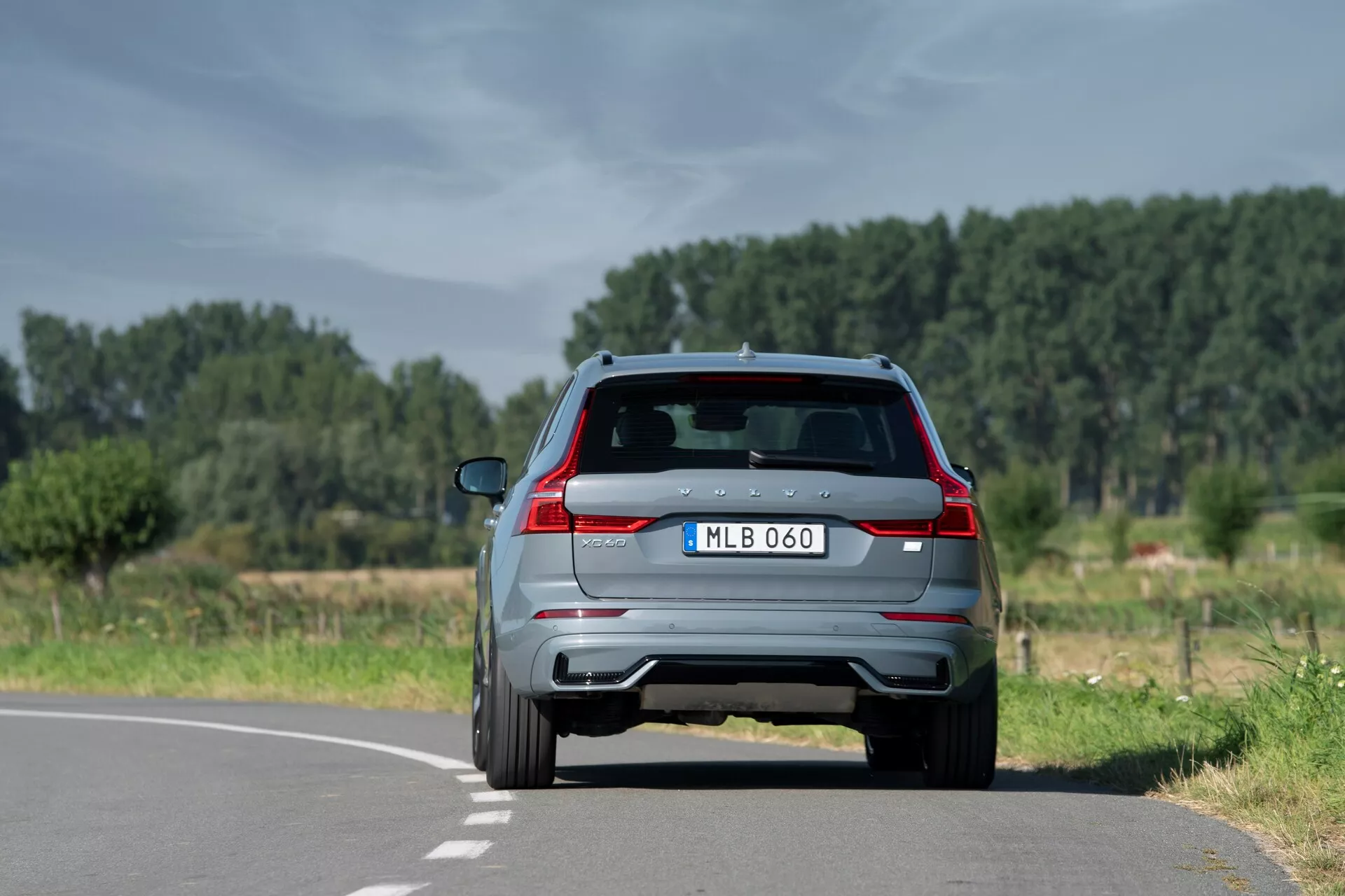 Vue arrière d'un SUV moderne Volvo XC60 2021 (2e génération-restylage) sur une route à deux voies dans un cadre naturel et rural, montrant des feux arrière distinctifs, une double sortie d'échappement, un aileron arrière avec feux de freinage intégrés, une caméra de recul, un essuie-glace arrière et des capteurs de stationnement. Positionné légèrement à droite de la ligne centrale, avec un ciel dégagé et des arbres en arrière-plan.
