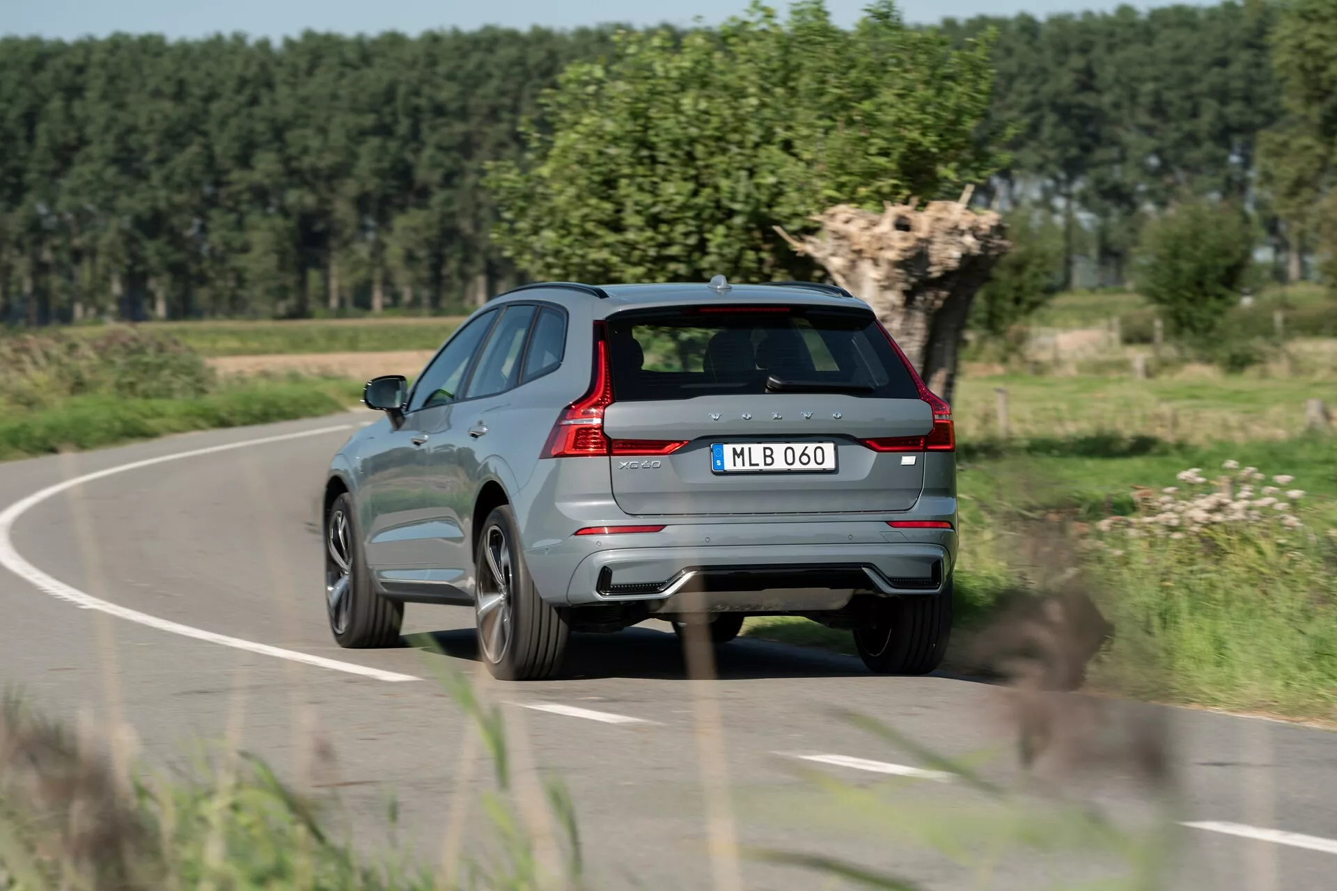 Vue arrière trois quarts d’un SUV Volvo XC60 2021 gris métallique moderne en mouvement sur une route de campagne, montrant des feux arrière distinctifs, un système d'échappement double, un essuie-glace arrière, un grand becquet arrière, des capteurs intégrés au pare-chocs arrière, une plaque d'immatriculation centrée, des jantes en alliage, une ligne de caractère marquée sur le côté, des garnitures de fenêtres réfléchissantes et un système de rails de toit. Arrière-plan avec feuillage flou et ciel dégagé indiquant le mouvement et un cadre rural.