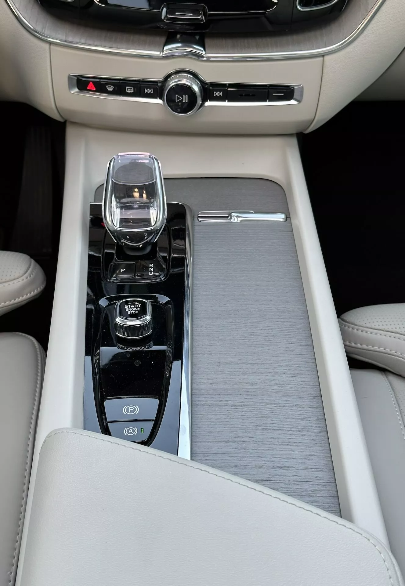 Vue rapprochée de la console centrale dans une Volvo XC60 recharge hybride plug-in 2021, dotée d'un accoudoir en cuir beige, d'un ornement coulissant en métal brossé, d'un contrôleur d'infodivertissement moderne, d'un sélecteur de vitesse en verre cristallin pour une transmission automatique, et de commandes pour le démarrage/arrêt du moteur, la désactivation de la traction et le mode de dynamique de conduite, ainsi qu'un contrôle de climatisation bi-zone.