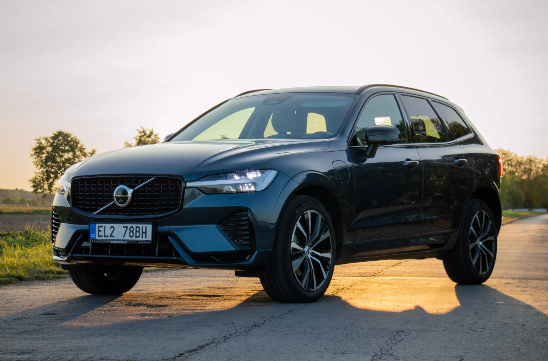 SUV moderne noir Volvo XC60 Recharge Plug-in Hybrid 2021 garé sur une route en asphalte au lever ou coucher du soleil, présentant son design avec un accent sur la grille, phares LED, jantes en alliage, et profil latéral sur fond de ciel en dégradé