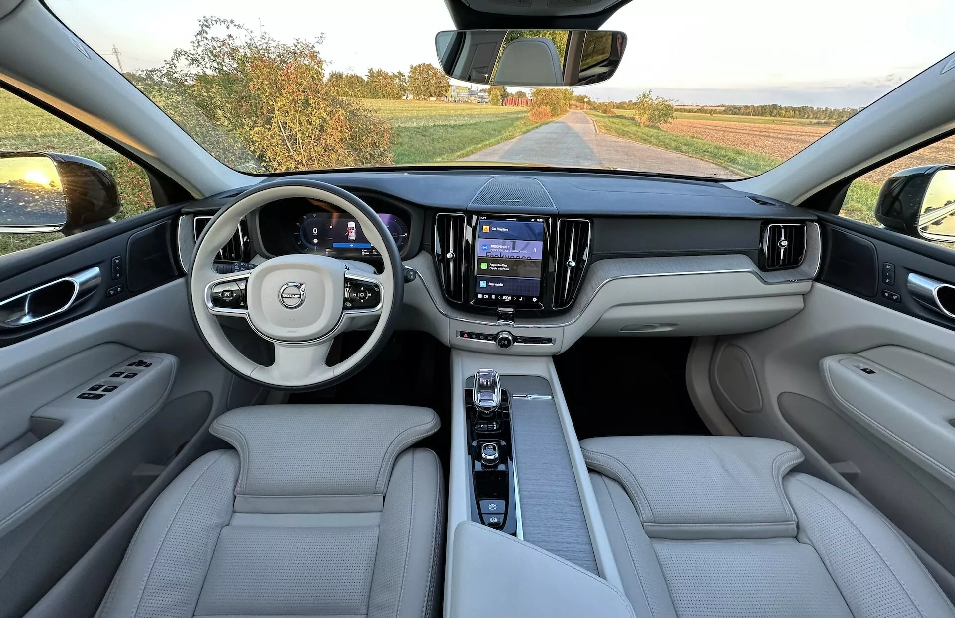 Vue intérieure d'une Volvo XC60 Recharge Plug-In Hybrid 2021 prise depuis la zone des sièges arrière montrant le tableau de bord complet, volant multifonction avec logo, tableau de bord numérique, système d'infodivertissement central tactile et sièges de luxe en cuir clair.