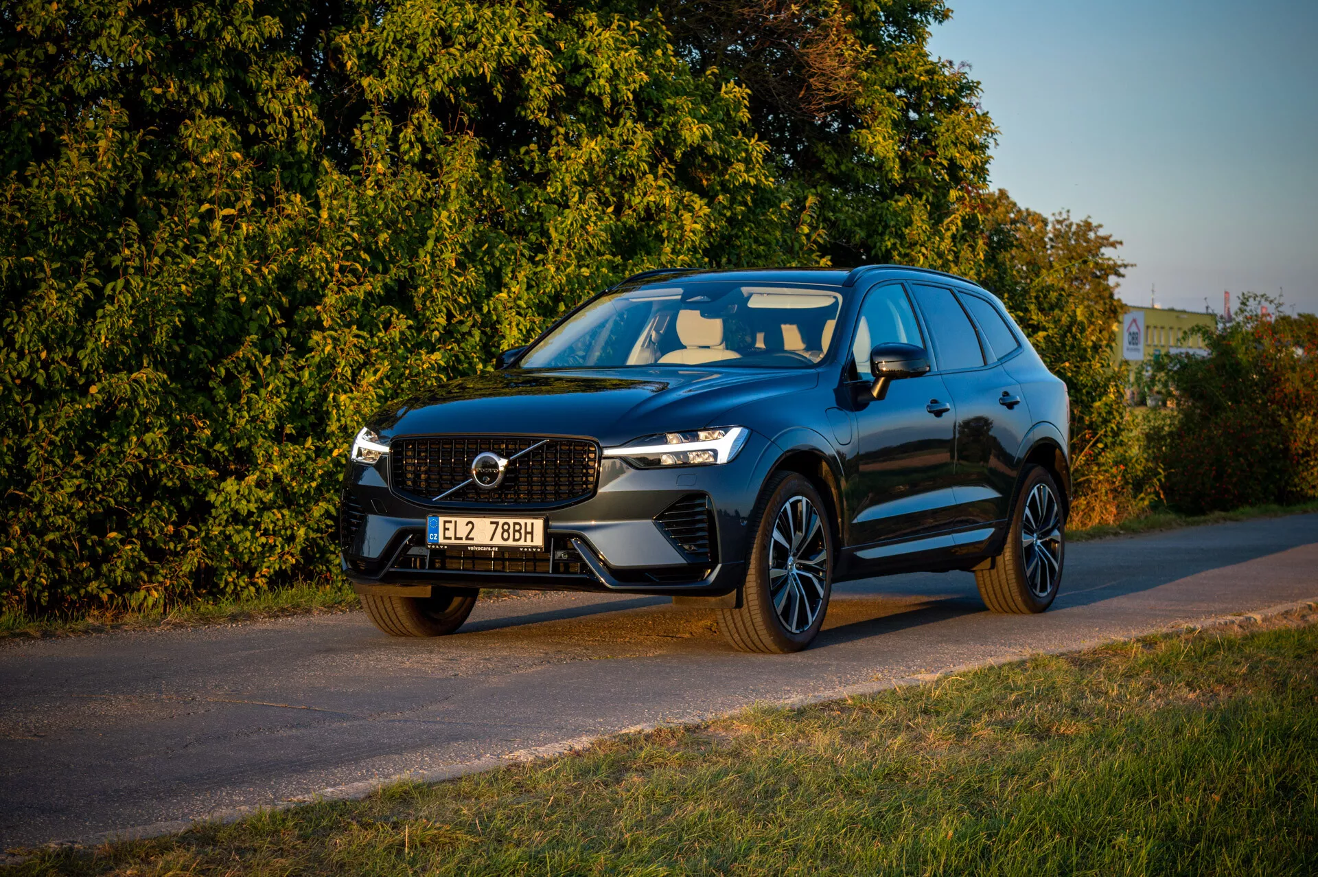 Volvo XC60 Recharge Plug-In Hybrid 2021 garée au bord de la route en fin d'après-midi avec un éclairage chaleureux, présentant la calandre avant, les phares LED et un profil sportif sur fond de végétation
