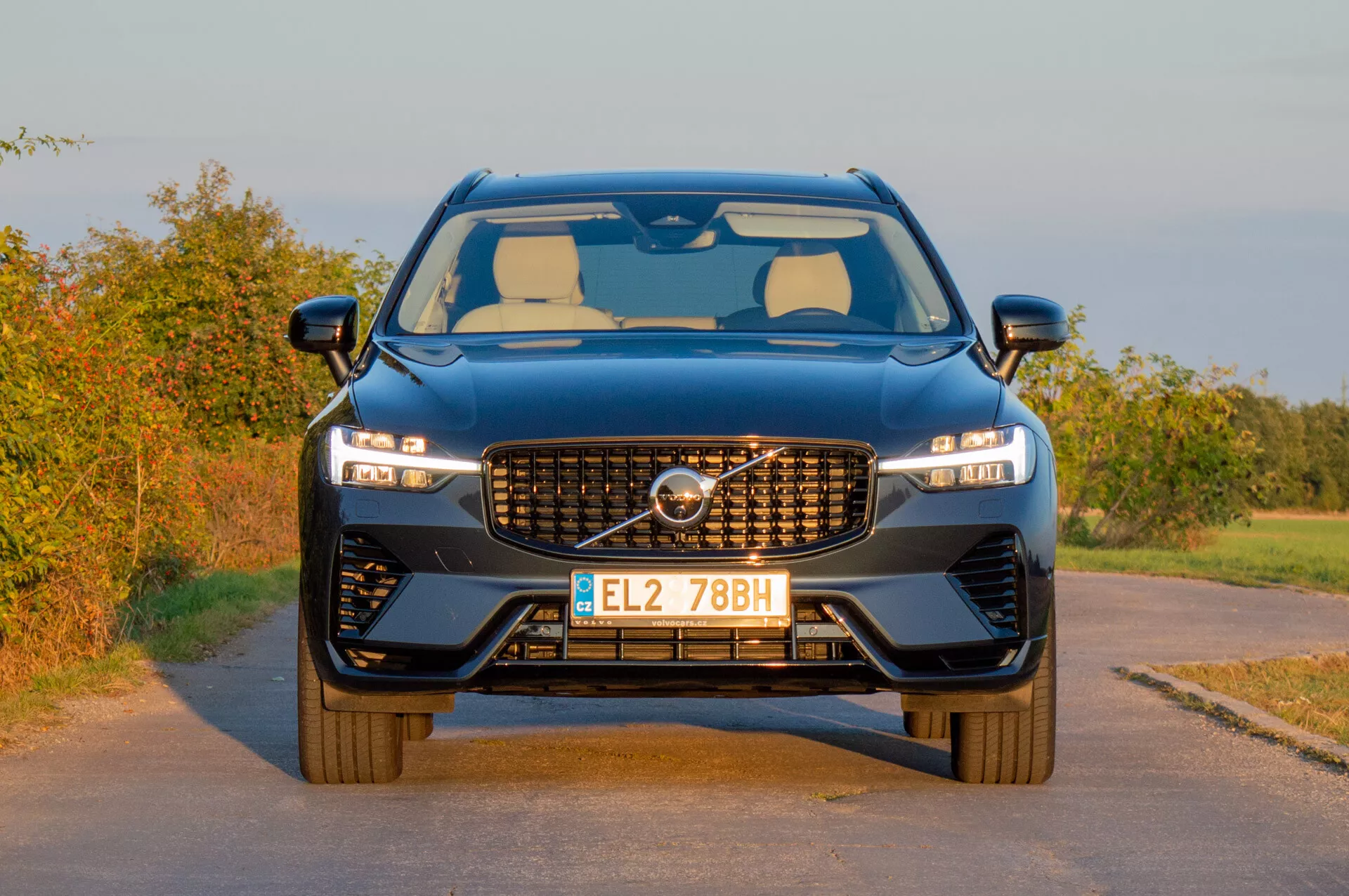 Volvo XC60 Recharge Plug-In Hybrid 2021 présentant son design moderne avec une grille distinctive, des phares LED élégants et un pare-chocs avant sportif sur fond rural dans une lumière chaude de fin d'après-midi