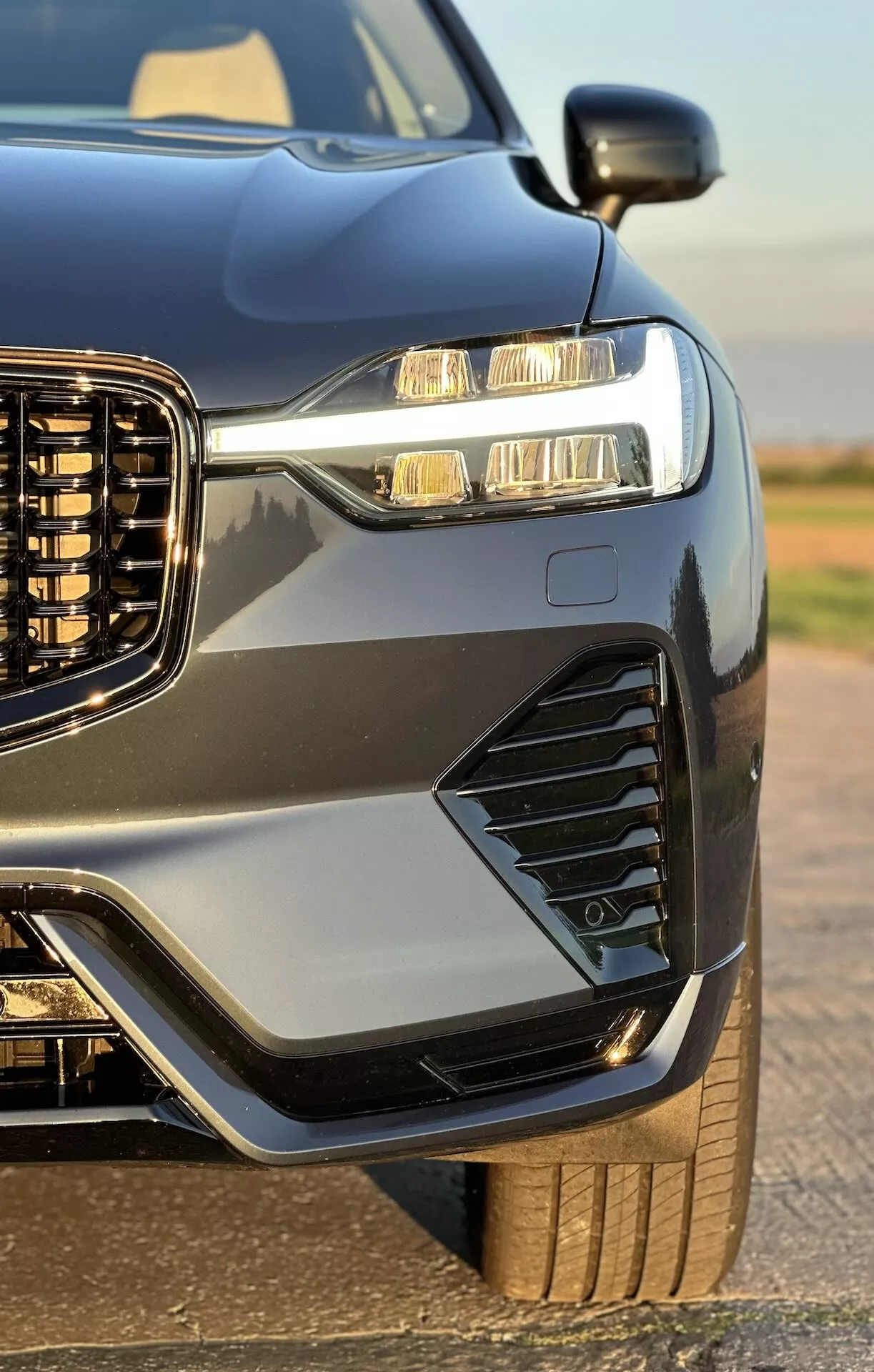 Vue rapprochée du quart avant d'une Volvo XC60 Recharge Hybride branchable 2021, mettant en lumière sa grille verticale, ses phares LED avec feux de jour, ses ventilations et feux de brouillard stylisés, son capot et ses ailes sportives, sa peinture métallique avec garnitures contrastantes, et un aperçu de sa roue robuste et de son pneu, rehaussé par la finition brillante du véhicule.