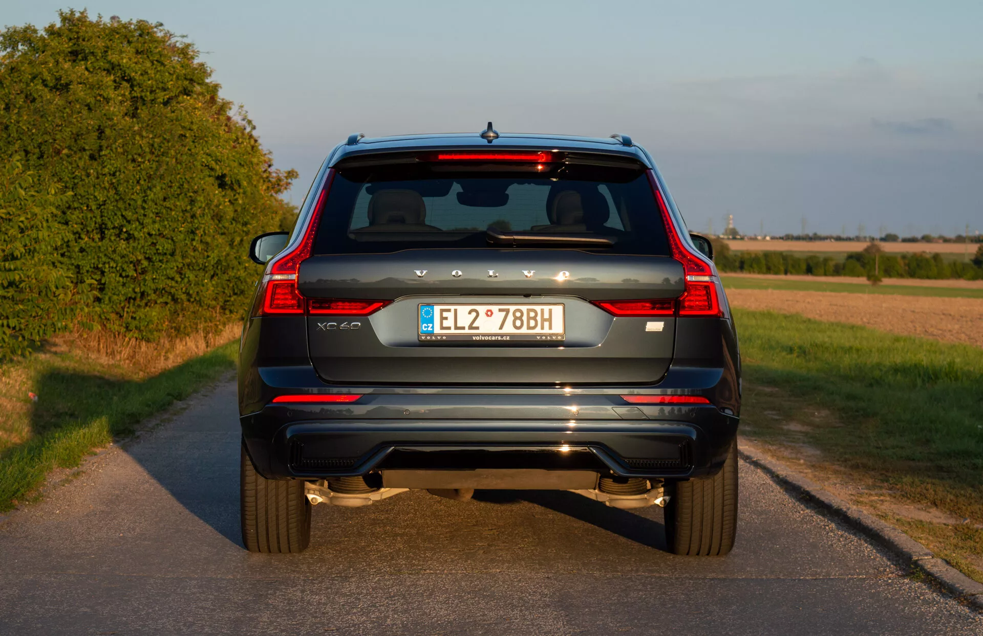 Vue arrière d'un SUV bleu Volvo XC60 Recharge Plug-In Hybrid 2021 avec feux arrière verticaux, embouts d'échappement doubles, réflecteurs arrière, essuie-glace arrière, troisième feu stop, plaque d'immatriculation européenne et caméra de recul sur une route rurale avant le coucher du soleil