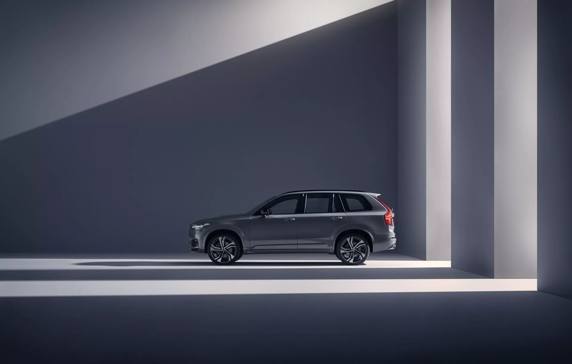 Profil latéral d'un Volvo XC90 2015 (2ème génération) de la catégorie SUV de luxe. La photo est prise dans un studio ou showroom intérieur, montrant les lignes de design du véhicule, des poignées de porte élégantes, de grandes roues en alliage multi-branches et des garnitures chromées sur les fenêtres. Les barres de toit suggèrent l'utilité en plus du luxe. Le véhicule est bien éclairé avec un éclairage dramatique créant des ombres géométriques, soulignant son apparence moderne et haut de gamme. Les caractéristiques avancées comme l'entrée sans clé sont suggérées par des éléments de design.