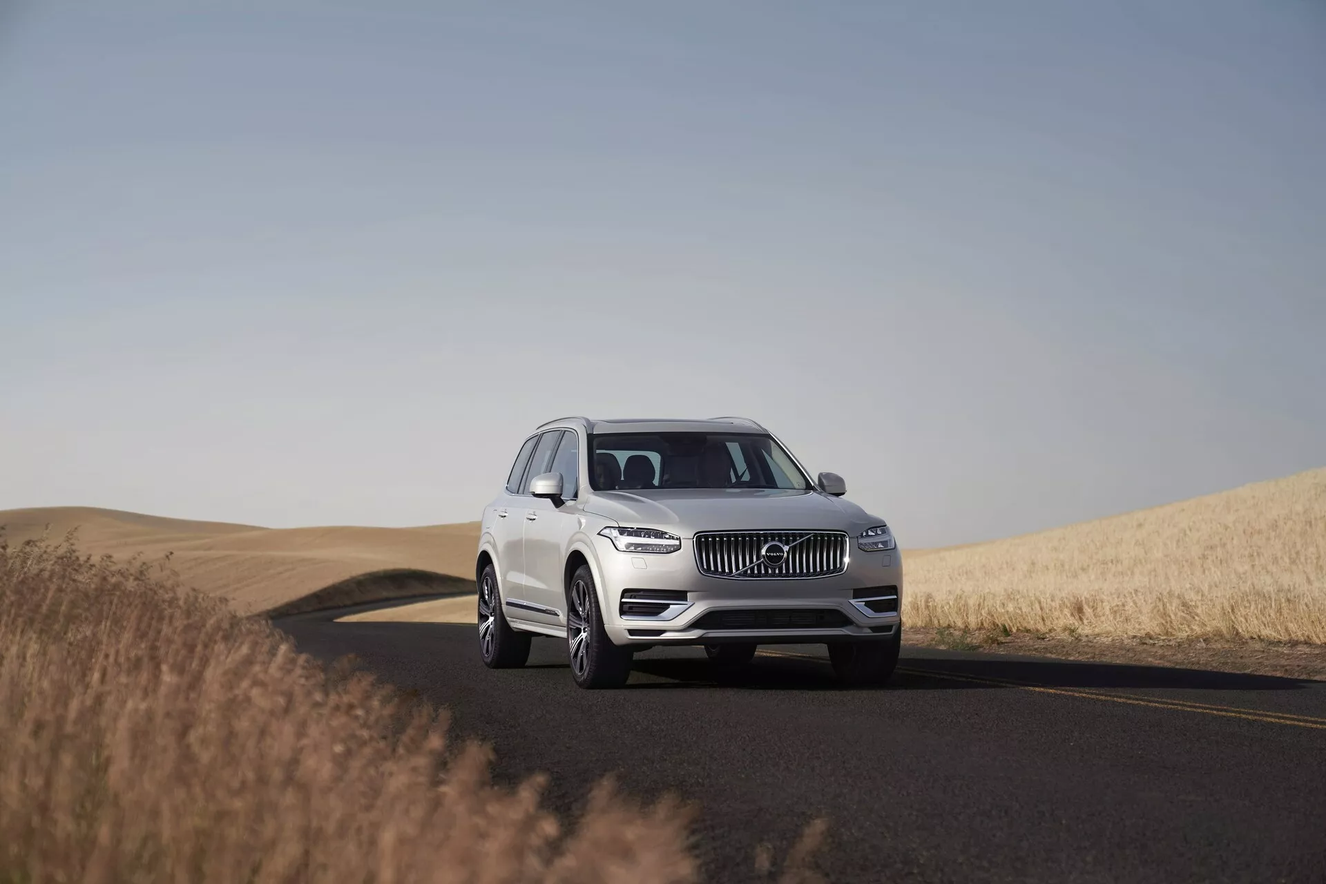 Un SUV Volvo XC90 blanc de 2015 sur une route à deux voies, capturé en plein jour sous un ciel clair. L'image montre le véhicule sous un angle bas à moyen, mettant en évidence sa présence imposante et ses dimensions. Positionné au centre du cadre avec une vue trois-quarts, la photo capture des détails de conception tels que la calandre, les phares LED, l'emblème et le pare-chocs avant, indiquant des systèmes avancés d'assistance à la conduite. Les roues, les alliages et éventuellement la taille des pneus sont clairement visibles. L'environnement serein présente une route qui tourne à droite et des champs de blé doré, soulignant l'aptitude du véhicule aux longs trajets dans des paysages divers.
