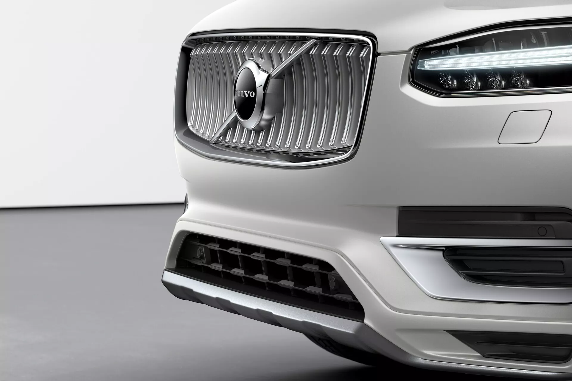 Gros plan sur la section avant d'une Volvo XC90 blanche de deuxième génération 2015, mettant en évidence sa calandre proéminente avec des lames verticales et l'emblème distinctif Volvo au centre. La finition chromée de la calandre contraste avec la texture mate qui l'entoure. Les feux de jour à LED Marteau de Thor sont visibles à l'intérieur du bloc optique. Le pare-chocs avant comporte une grande prise d'air soulignée par une garniture argentée, suggérant une efficacité aérodynamique et un refroidissement du moteur. Pris sous un angle légèrement inférieur, l'image met en valeur l'élégance, la qualité de fabrication et le design premium de l'avant de Volvo.