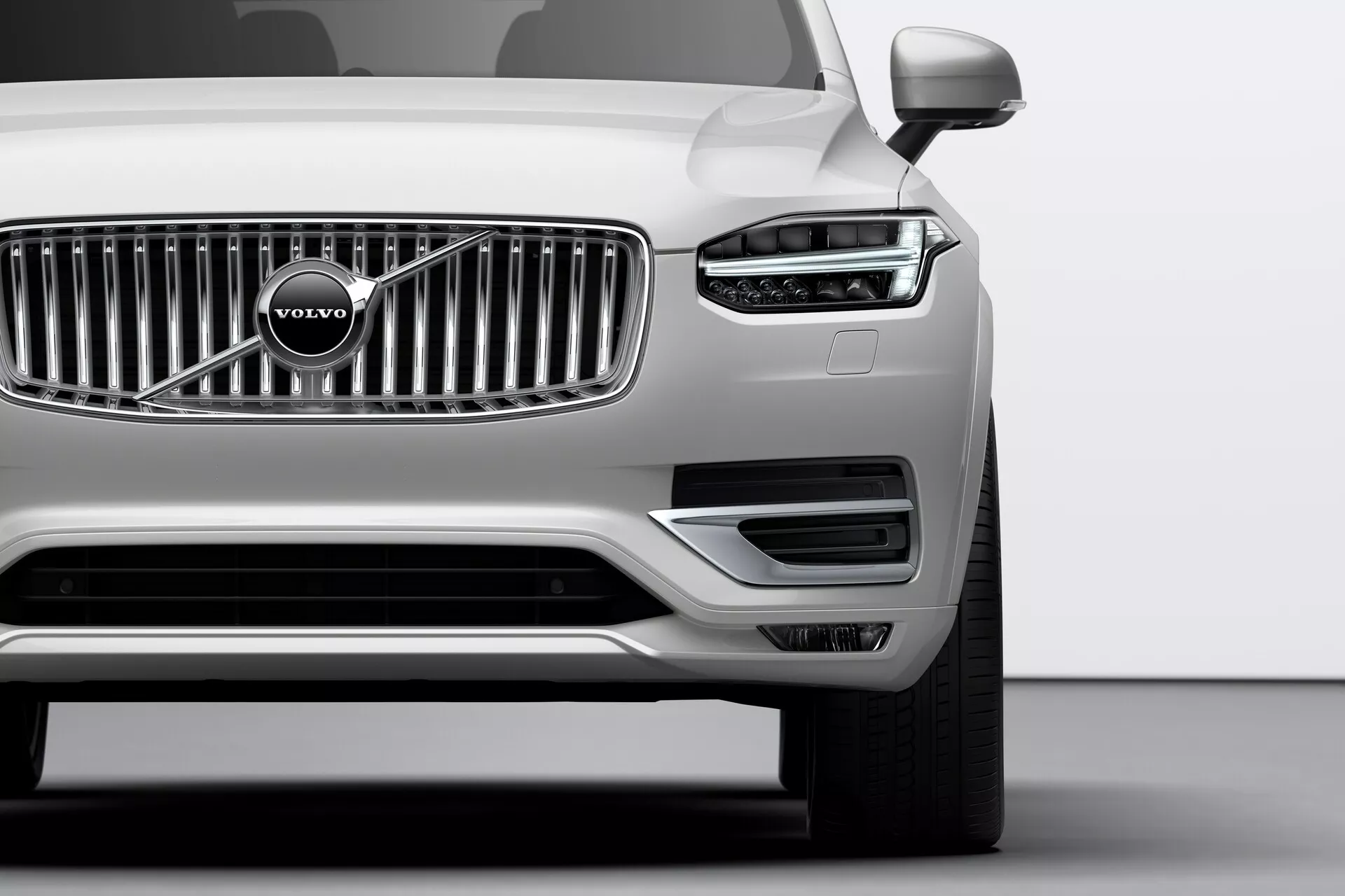 Gros plan sur le quart avant d'un Volvo XC90 argenté de 2015 (2ème génération) pris sous un angle bas. On remarque la grille chromée proéminente avec l'emblème emblématique de Volvo, les phares élégants avec des feux diurnes à LED en forme de T, appelés 'Marteau de Thor', et les capteurs de stationnement bien intégrés dans le pare-chocs avant. La partie inférieure du pare-chocs comprend des prises d'air et des logements pour les phares antibrouillard encadrés par une garniture argentée. Les rétroviseurs extérieurs visibles laissent entrevoir des caractéristiques technologiques avancées telles que des répétiteurs de clignotants ou des systèmes d'alerte d'angle mort.