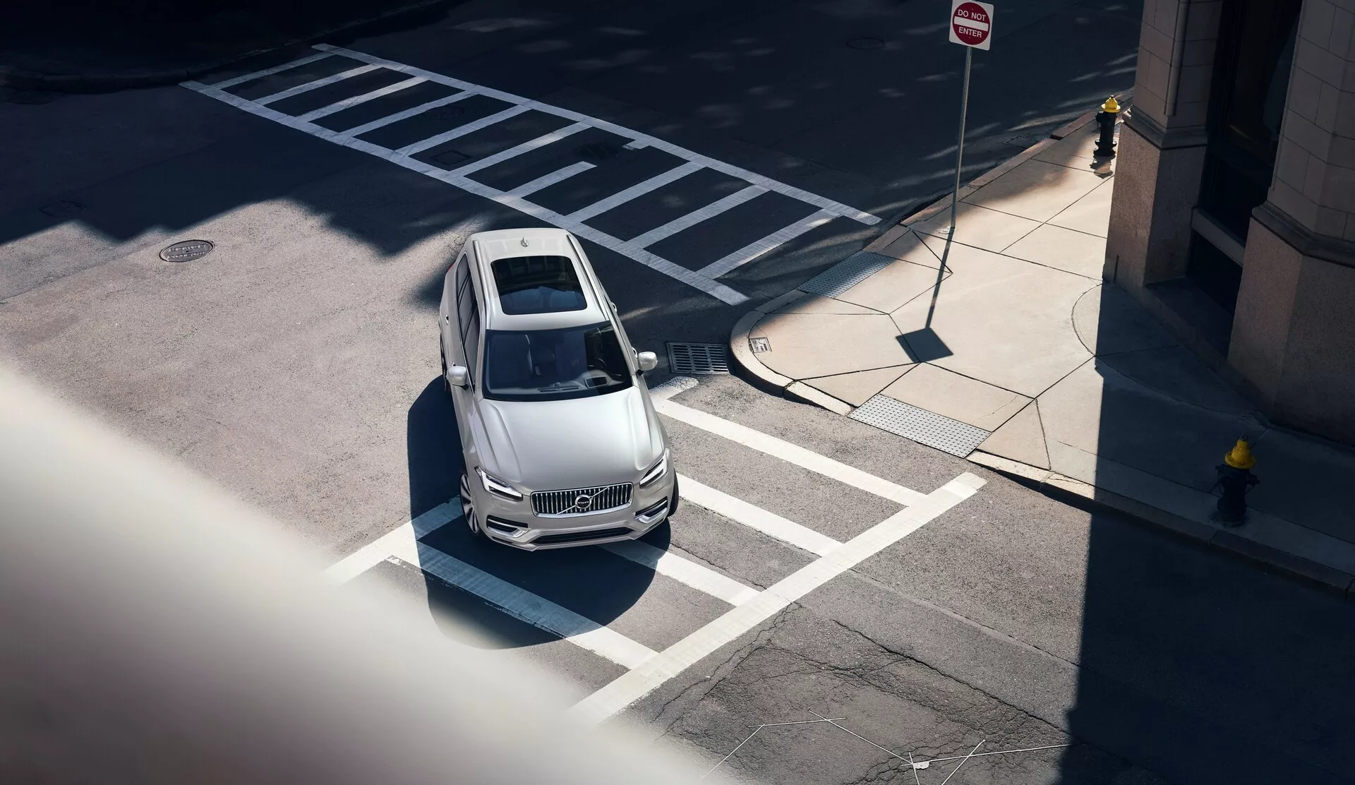 Vue aérienne d'une Volvo XC90 2015 (2ème génération) depuis un angle élevé, vue en diagonale depuis le coin avant gauche; montrant le capot clair, le toit et une partie du côté conducteur. Le véhicule, correctement positionné dans une place de parking marquée près d'un passage piéton, présente des lignes de design indiquant une aérodynamique, un pare-brise clair révélant un intérieur partiellement visible, un toit ouvrant notable et des lignes latérales nettes. Le cadre urbain est accentué par les panneaux de signalisation de stationnement et de circulation à proximité, avec des ombres marquées suggérant une lumière de midi. Le véhicule laisse aussi entrevoir des caractéristiques technologiques modernes sans capteurs ni caméras visibles.