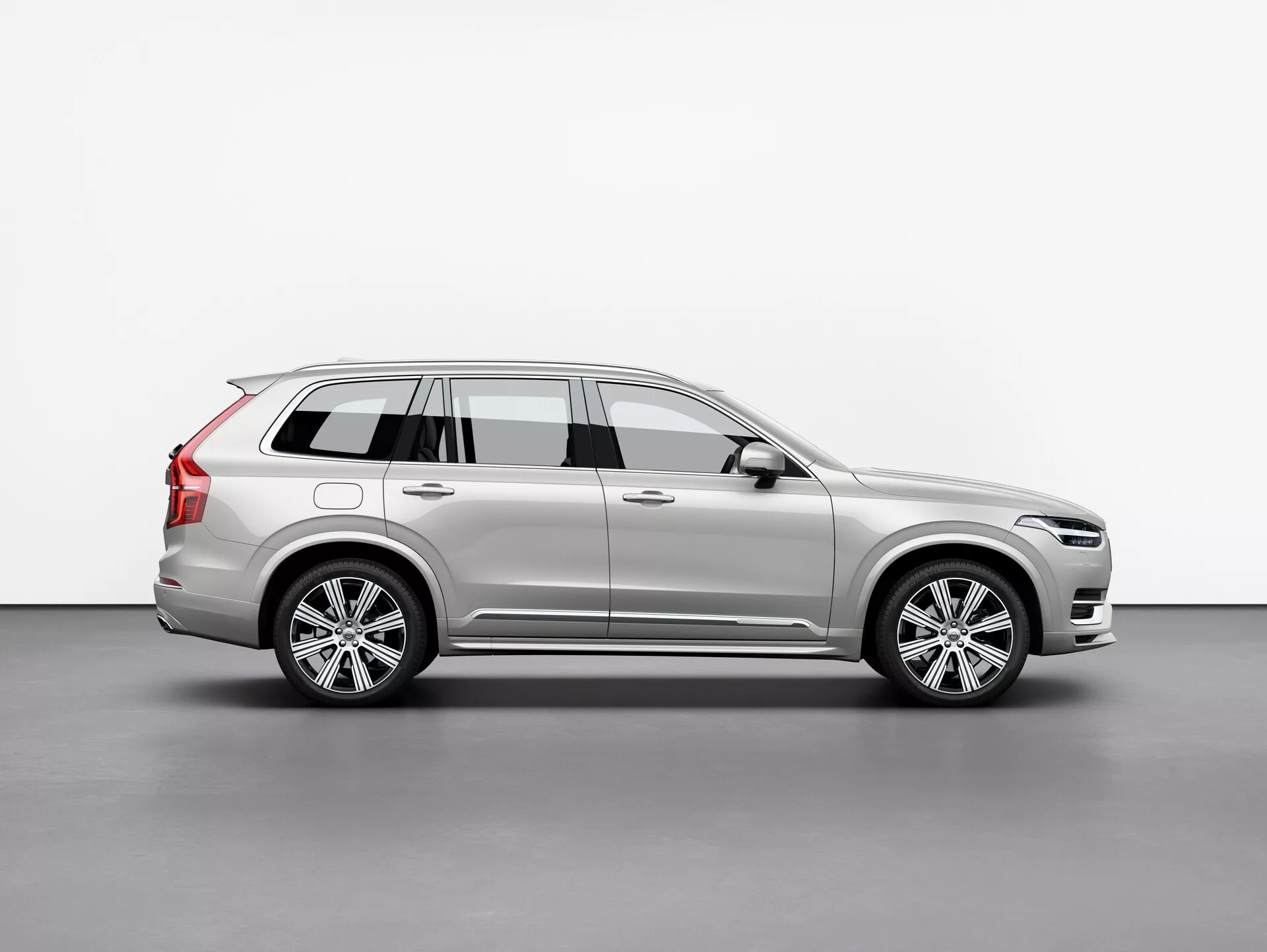 Image de profil latéral d'un SUV Volvo XC90 2015 (2ème génération) argenté, exhibant ses roues en alliage, ses barres de toit, son rétroviseur latéral avec signal de virage intégré et les lignes détaillées de sa carrosserie, positionné au centre sur un fond simple.