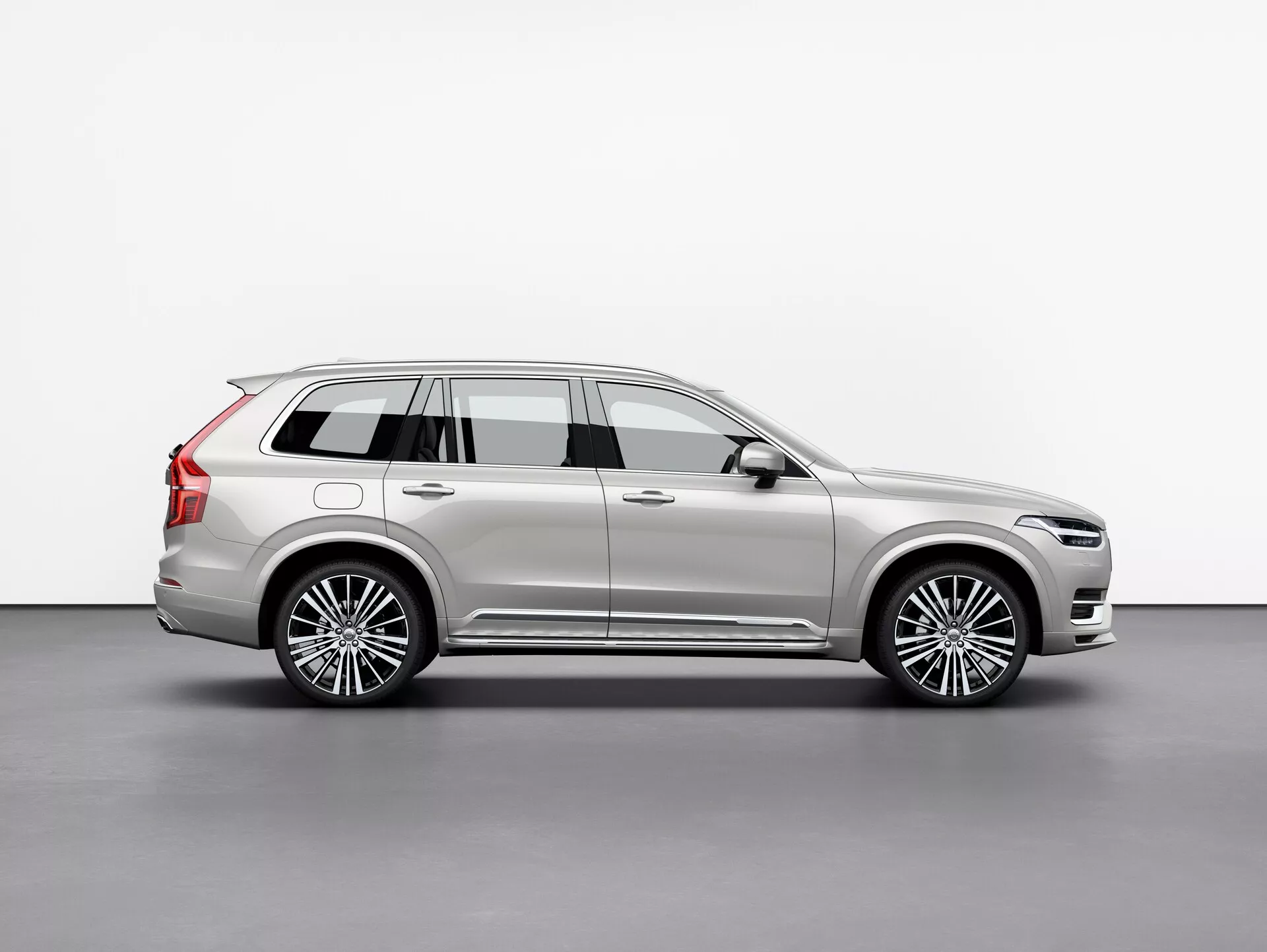 Profil latéral de la Volvo XC90 2015, un SUV de luxe de grande taille, capturé dans un studio avec un fond blanc uni. L'image met en évidence l'empattement long du véhicule, les quatre portes, le hayon arrière, les roues en alliage à rayons multiples, les garnitures chromées, la peinture métallique et les éléments de design proéminents tels que les rétroviseurs latéraux et les barres de toit. La scène soigneusement éclairée met en valeur les lignes et les courbes de la carrosserie, reflétant sa nature haut de gamme. Le véhicule est positionné pour offrir une vue claire de sa longueur, de sa hauteur et de sa posture globale, indicatif d'un SUV à trois rangées pour passagers et chargement.