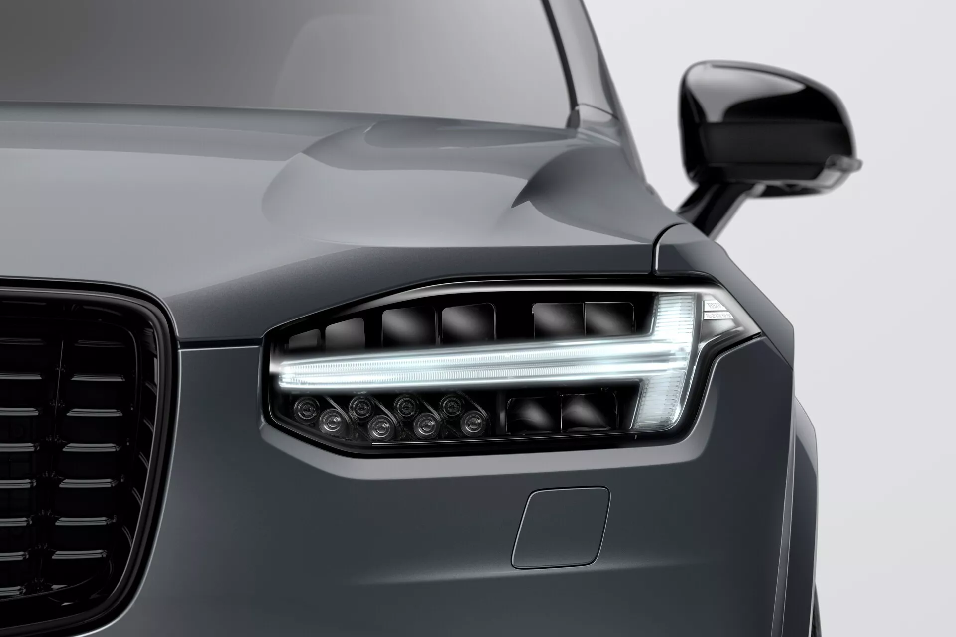 Gros plan sur l'avant d'une Volvo XC90 2015 (2ème génération), mettant en valeur le phare gauche avec une technologie LED ou laser moderne, des éléments d'éclairage doubles, des feux de jour élégants et plusieurs lentilles de projecteur pour une visibilité améliorée. L'image met en évidence la grande calandre esthétiquement conçue, le rétroviseur latéral aérodynamique de la couleur de la carrosserie et le capteur ADAS potentiel sous le phare. La finition lisse et réfléchissante de couleur gris métallisé profond met en avant les lignes élégantes mais musclées du capot et de l'aile, démontrant une peinture de haute qualité et un design de véhicule de pointe.