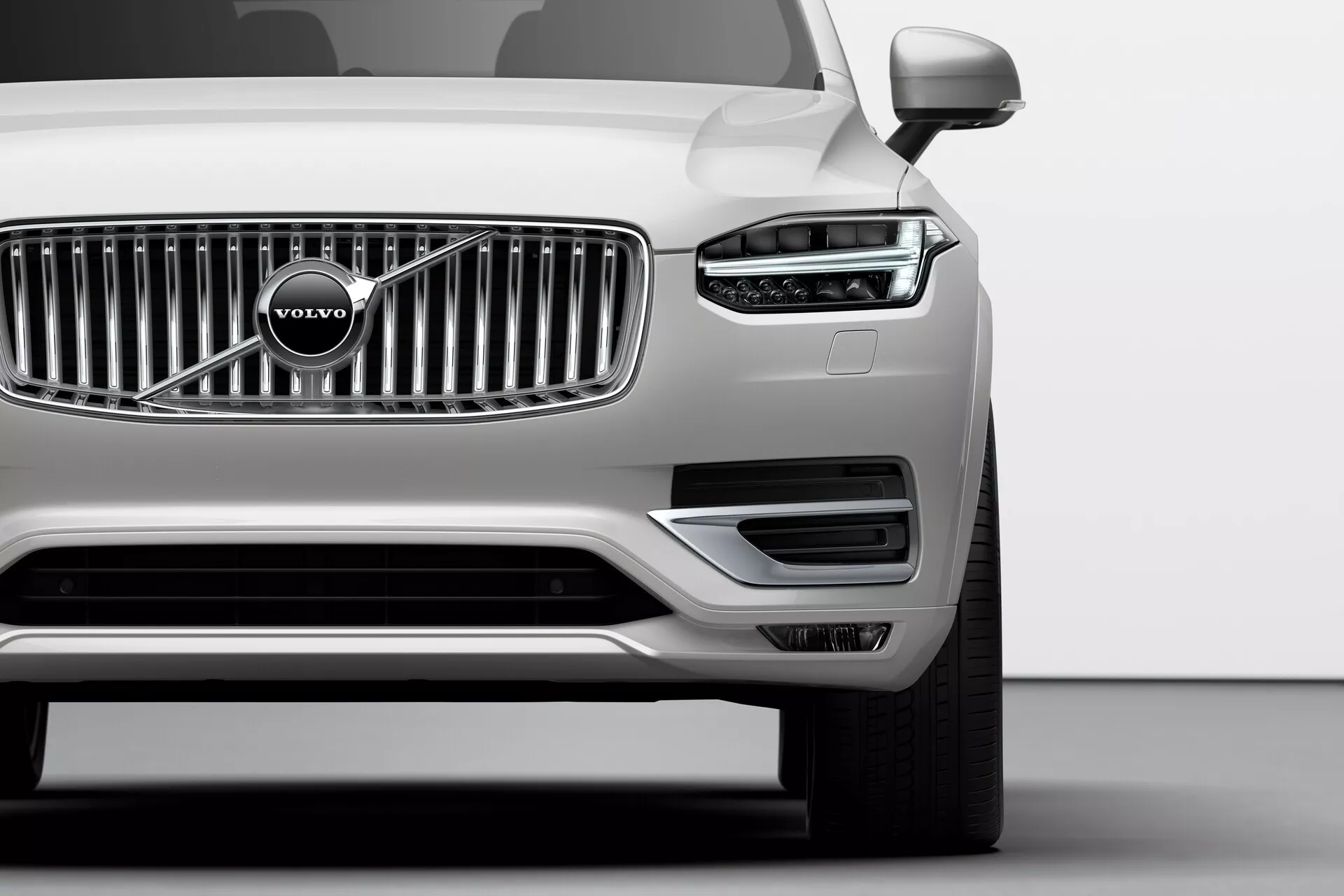 Prise en angle bas du quart avant gauche d'une Volvo XC90 argentée de 2015 (2e génération) dans un studio, mettant en avant le motif distinctif en forme de marteau de Thor de la lumière diurne LED, la calandre à lames chromées verticales avec l'emblème Volvo, le pare-chocs avant minimaliste avec des prises d'air, le rétroviseur latéral gauche, la roue en alliage multi-brins et la finition métallique impeccable, illustrant la technologie avancée de l'éclairage, le style de la marque et les caractéristiques aérodynamiques.