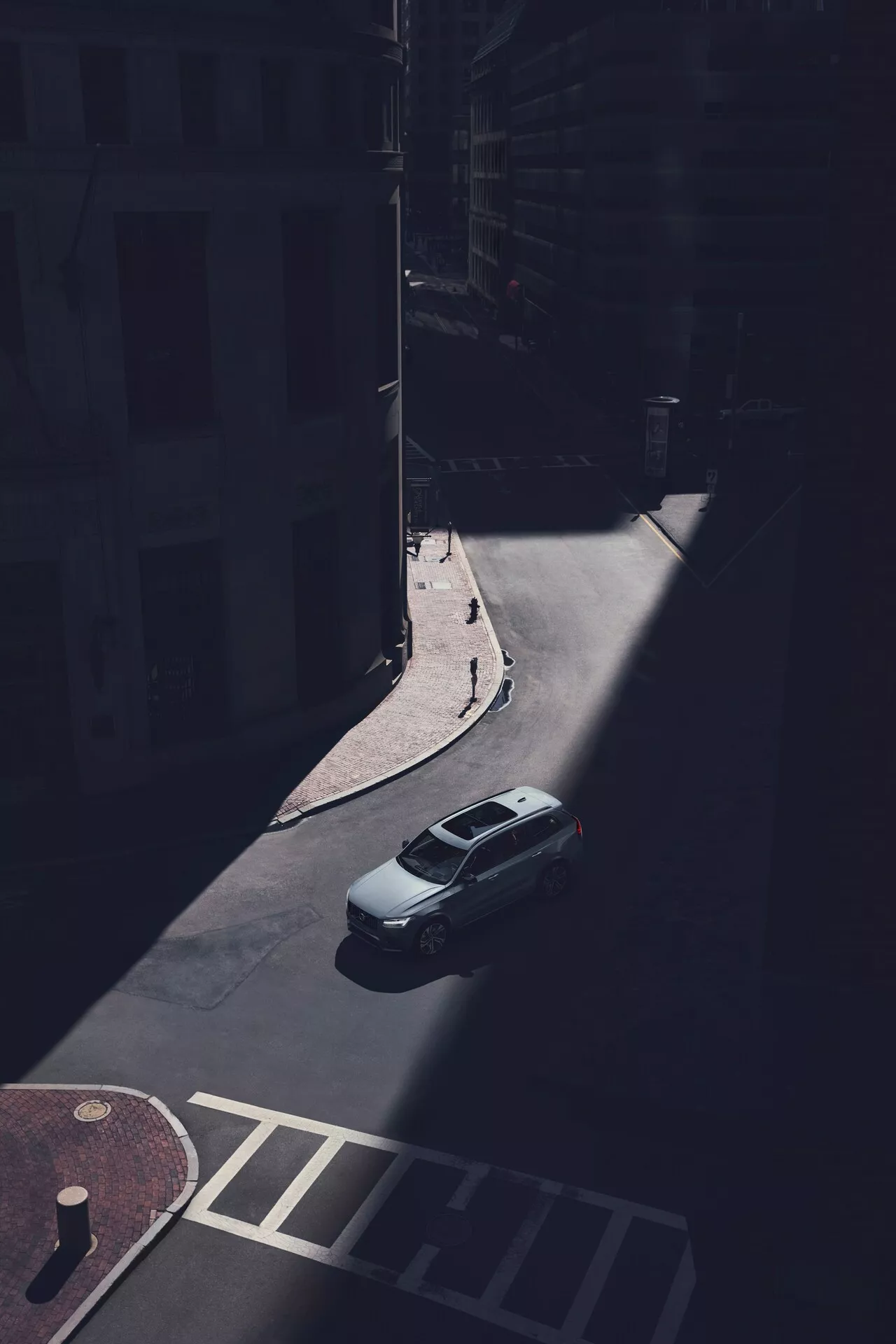 Vue aérienne de la berline Volvo XC90 2015 (2ème génération) stationnée dans une rue urbaine ombragée, capturée sous un angle oblique depuis un point de vue élevé. Le coffre, le toit et la lunette arrière de la voiture sont clairement visibles, avec l'arrière du véhicule illuminé par la lumière du soleil. L'image capture le couvercle du coffre arrière, la forme de la lunette arrière et la partie supérieure du coffre, tandis que les ombres dissimulent les détails spécifiques du design, comme les feux arrière et le pare-chocs. La ligne de toit aérodynamique est évidente, sans barres de toit ni toits ouvrants visibles. La voiture est stationnée le long d'un trottoir près des façades des bâtiments et d'un passage piéton, mettant en évidence l'isolement du véhicule dans l'environnement urbain ombragé.