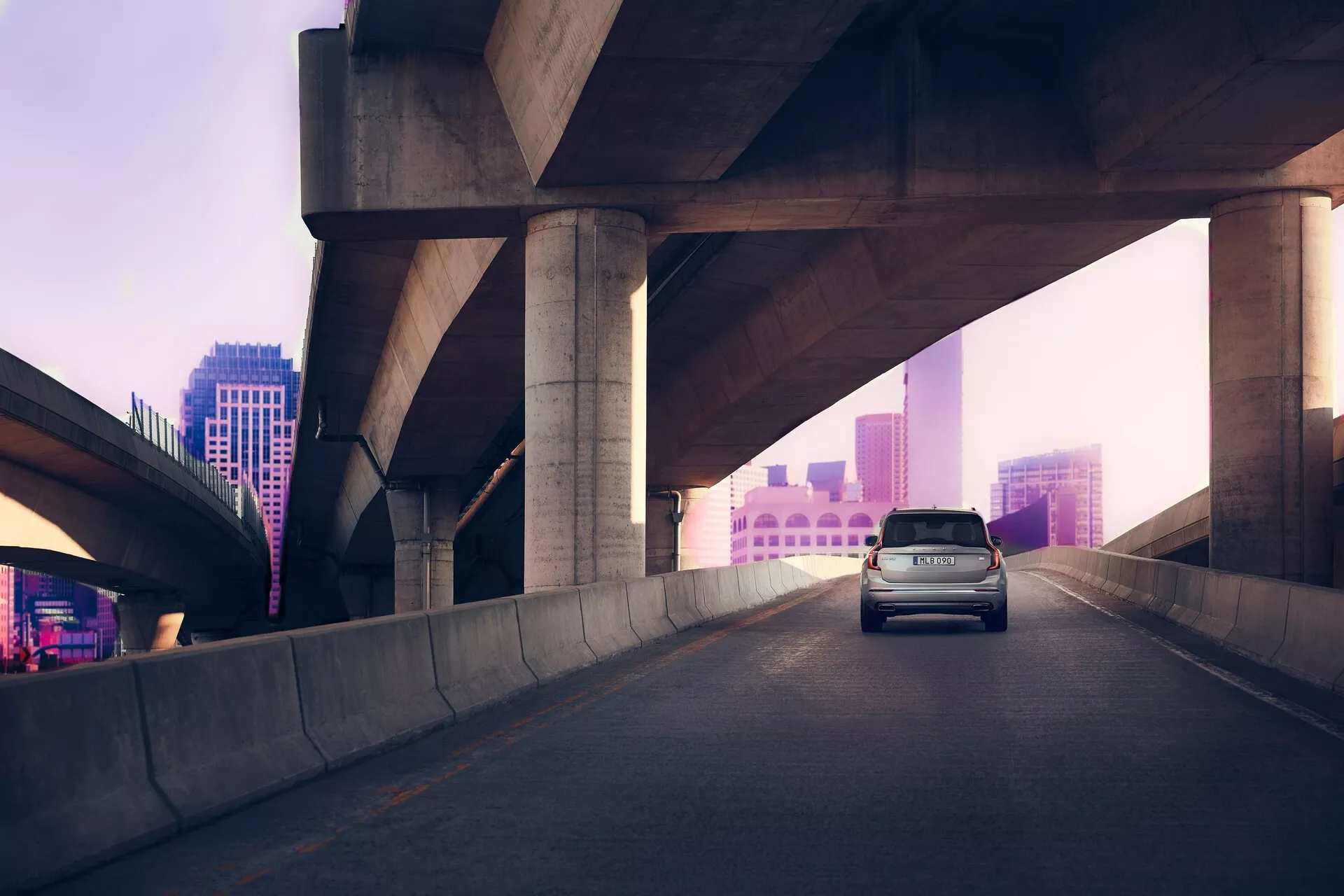 Vue trois quarts arrière d'une Volvo XC90 de deuxième génération 2015 sur une autoroute surélevée, avec en arrière-plan une ville éclairée en rose-violet à l'aube ou au crépuscule. Les feux de freinage du véhicule sont allumés, suggérant une décélération. Le SUV est doté d'une ligne de toit haute, d'un hayon et d'un design aérodynamique pour l'efficacité et le style, capturé en lumière naturelle diffuse.