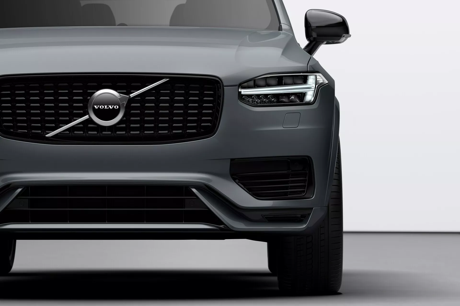 Vue en gros plan de l'angle avant de la Volvo XC90 2015 (2e génération) centrée sur le côté gauche, mettant en avant la calandre distinctive, le grand emblème Volvo et le design des phares à LED 'Marteau de Thor'. L'image comprend le phare, la roue avant, une partie de la porte du conducteur, des capteurs visibles sur le pare-chocs inférieur et le rétroviseur latéral avec une possible technologie de surveillance des angles morts.