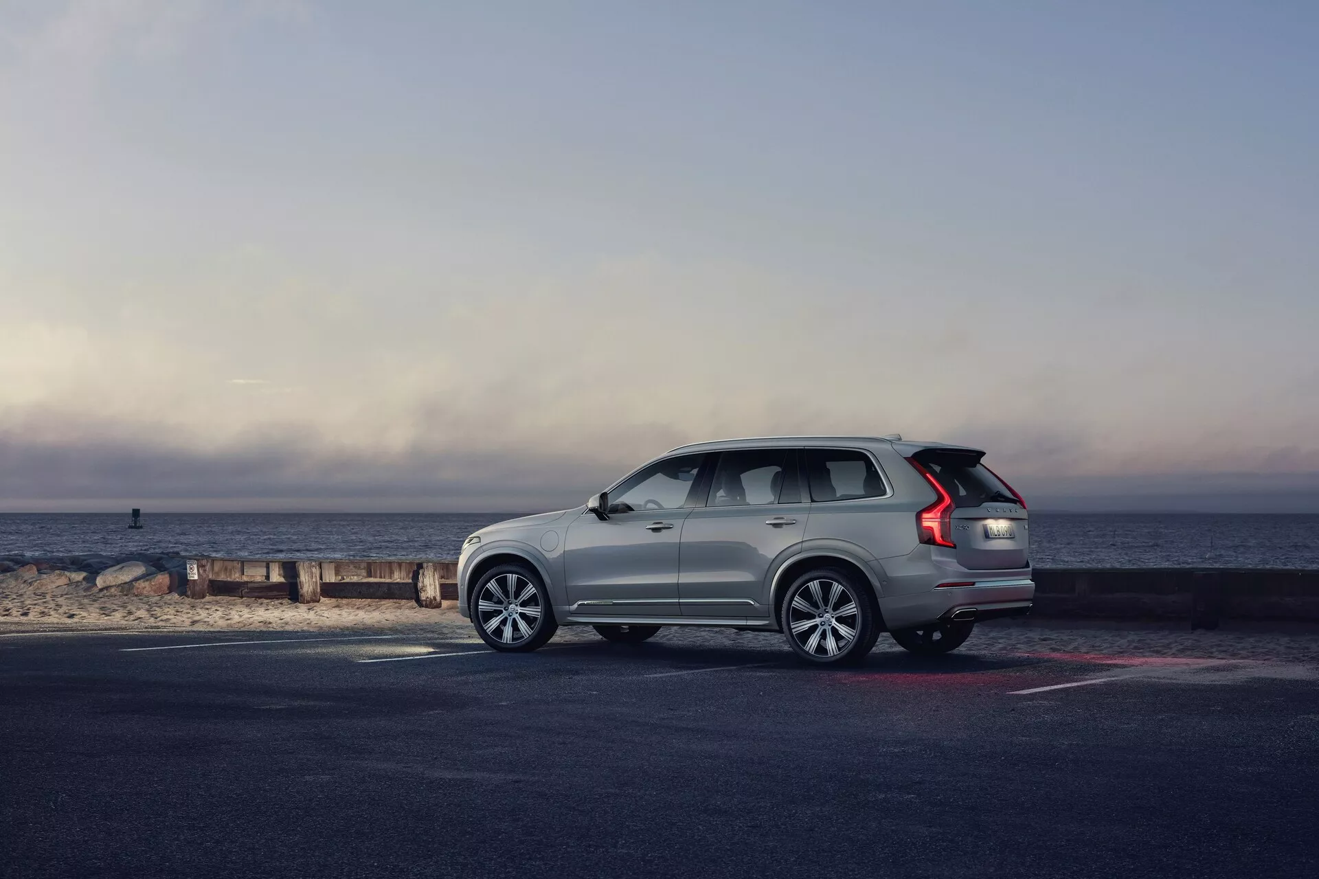 Vue de profil d'un SUV Volvo XC90 2015 (2e génération) garé au bord de la mer, le véhicule étant orienté vers la droite. L'angle met en avant le côté et l'arrière, montrant des éléments de design tels que les roues en alliage multibrins, les feux arrière à LED allongés, les barres de toit, la ligne de fenêtre qui monte vers l'arrière et les poignées de porte affleurantes. Le SUV de couleur claire se détache sur le pavé plus sombre et la plage, baigné par la lumière naturelle du matin ou de fin d'après-midi, avec un fond pittoresque de l'horizon où le ciel rencontre la mer. La composition générale évoque un sentiment d'aventure, de luxe et de liberté.