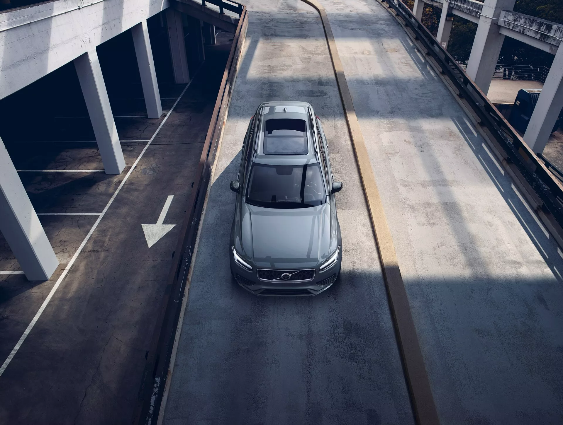 Vue aérienne d'une berline Volvo XC90 2015 grise ou argentée, circulant sur une route urbaine à plusieurs niveaux, montrant son toit panoramique, son design aérodynamique, ses phares LED élégants et ses grandes jantes en alliage, suggérant un véhicule moderne et luxueux.