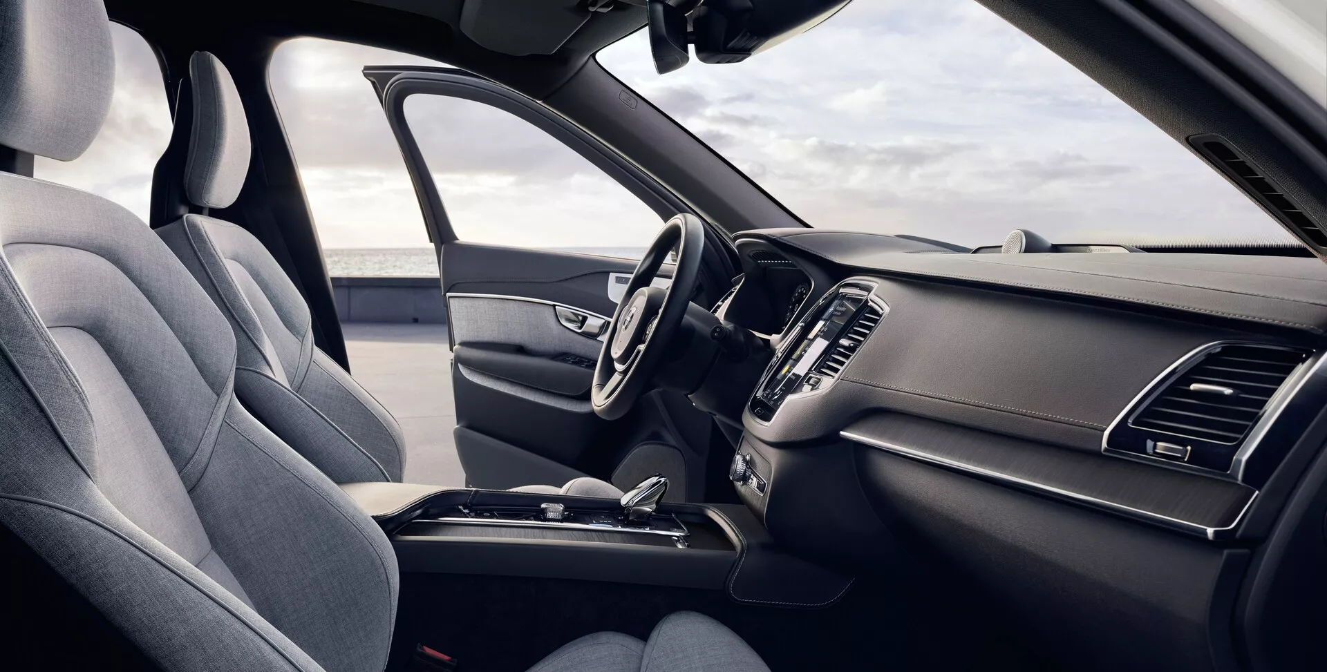 Cette image présente l'intérieur d'une Volvo XC90 de deuxième génération de 2015, vue du côté conducteur. L'angle légèrement surélevé offre une vue détaillée des sièges avant et du tableau de bord, avec un siège conducteur à gauche garni de tissu texturé, doté d'un appui-tête visible et d'un soutien latéral. Le volant multifonctionnel comporte plusieurs boutons de commande et le tableau de bord élégant comprend des aérateurs symétriques et un écran d'infodivertissement central partiellement visible. La console centrale montre un levier de vitesse automatique et d'autres caractéristiques de commande, soulignant un design ergonomique et pratique.