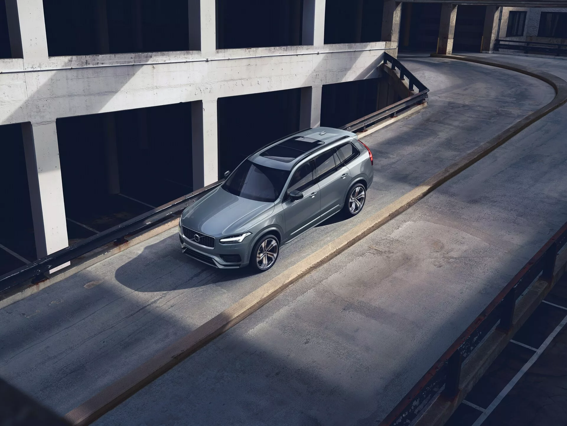 Vue en plongée d'un SUV Volvo XC90 2015 garé sur une surface en béton, mettant en valeur son design élégant et moderne avec une finition de peinture métallique, une calandre distinctive, des phares visibles et des roues en alliage complexes. L'angle de vue met en évidence le toit, le capot et le côté gauche du véhicule, avec une ombre suggérant une lumière matinale ou de fin d'après-midi. L'environnement semble être un niveau supérieur d'une structure de stationnement à plusieurs étages, mettant en valeur la prestance du véhicule dans un environnement urbain propre.