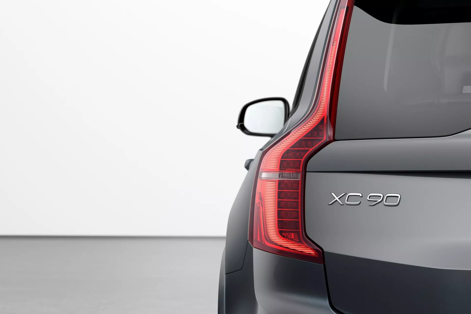 Vue rapprochée du coin arrière d'une Volvo XC90 2015 (2ème génération) mettant en valeur les détails du design : feu arrière LED vertical, nom du modèle 'XC90', pare-brise arrière élégant avec spoiler, et finition de peinture métallique premium sur un fond clair.