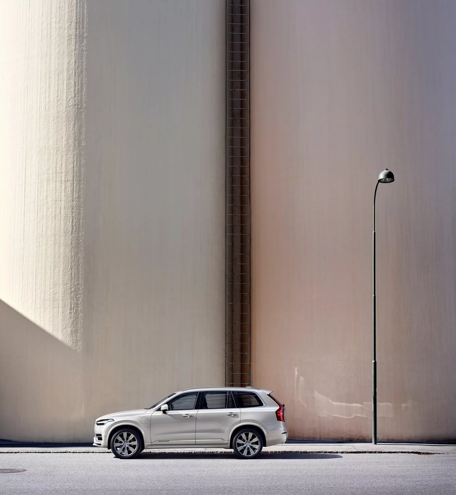 Vue de profil latéral d'un SUV Volvo XC90 2015 blanc garé parallèlement à un mur beige lisse et haut avec un tuyau vertical, avec un arrière-plan urbain comprenant un lampadaire. La voiture, positionnée à gauche du cadre avec les rétroviseurs déployés, projette une légère ombre indiquant la lumière naturelle du jour. L'image met également en avant des éléments de design moderne tels que des jantes en alliage et des phares LED, soulignant les caractéristiques automobiles clés.