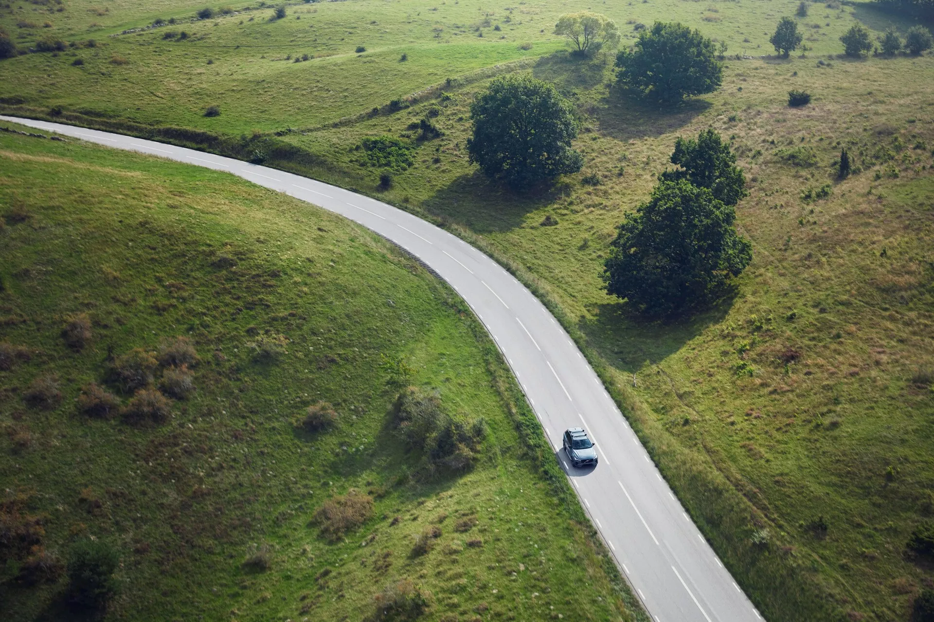 Vue aérienne d'un Volvo XC90 2015 argenté ou gris clair roulant sur une route rurale sinueuse entourée de champs verts avec de petits arbres et arbustes. La voiture, capturée d'une perspective en plongée, est positionnée au milieu de la courbe à droite du centre de l'image, projetant une ombre suggérant un soleil de midi.