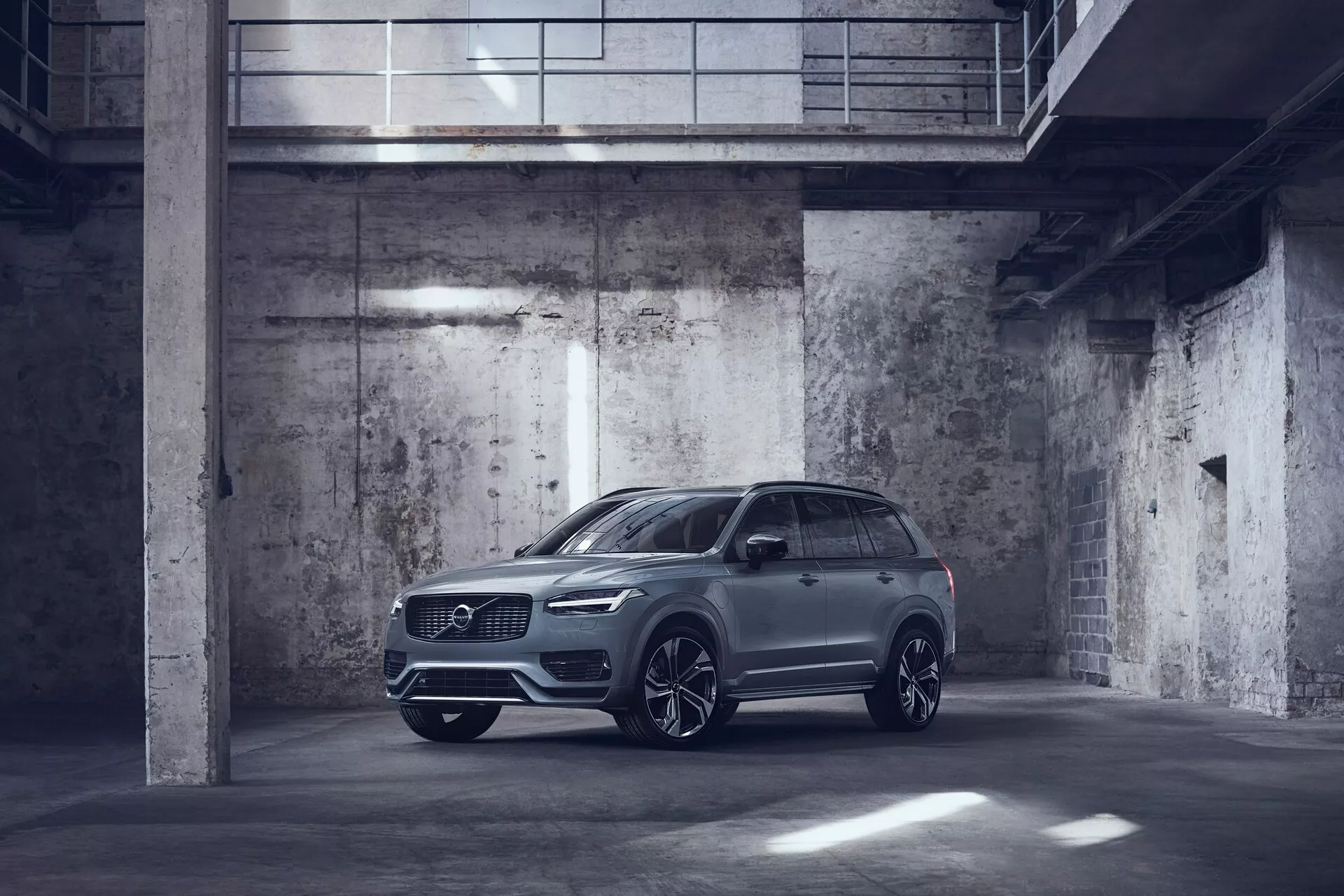 Image de la Volvo XC90 2015 de couleur métallique garée dans un parking de style industriel faiblement éclairé. Capturée sous un angle avant trois-quarts, montrant le côté conducteur avec la face avant et le profil latéral bien visibles. La voiture dispose de phares LED élégants, d'une grande calandre avant, possiblement de capteurs de stationnement et de grandes jantes en alliage avec un design à rayons multiples. Les rétroviseurs latéraux sont de forme aérodynamique et peuvent inclure des fonctionnalités technologiques avancées comme les indicateurs de signalisation et les systèmes d'alerte de l'angle mort. L'image met en évidence le design luxueux mais utilitaire du SUV avec une garde au sol élevée et des fonctionnalités potentielles sur le toit, comme un toit ouvrant ou panoramique.