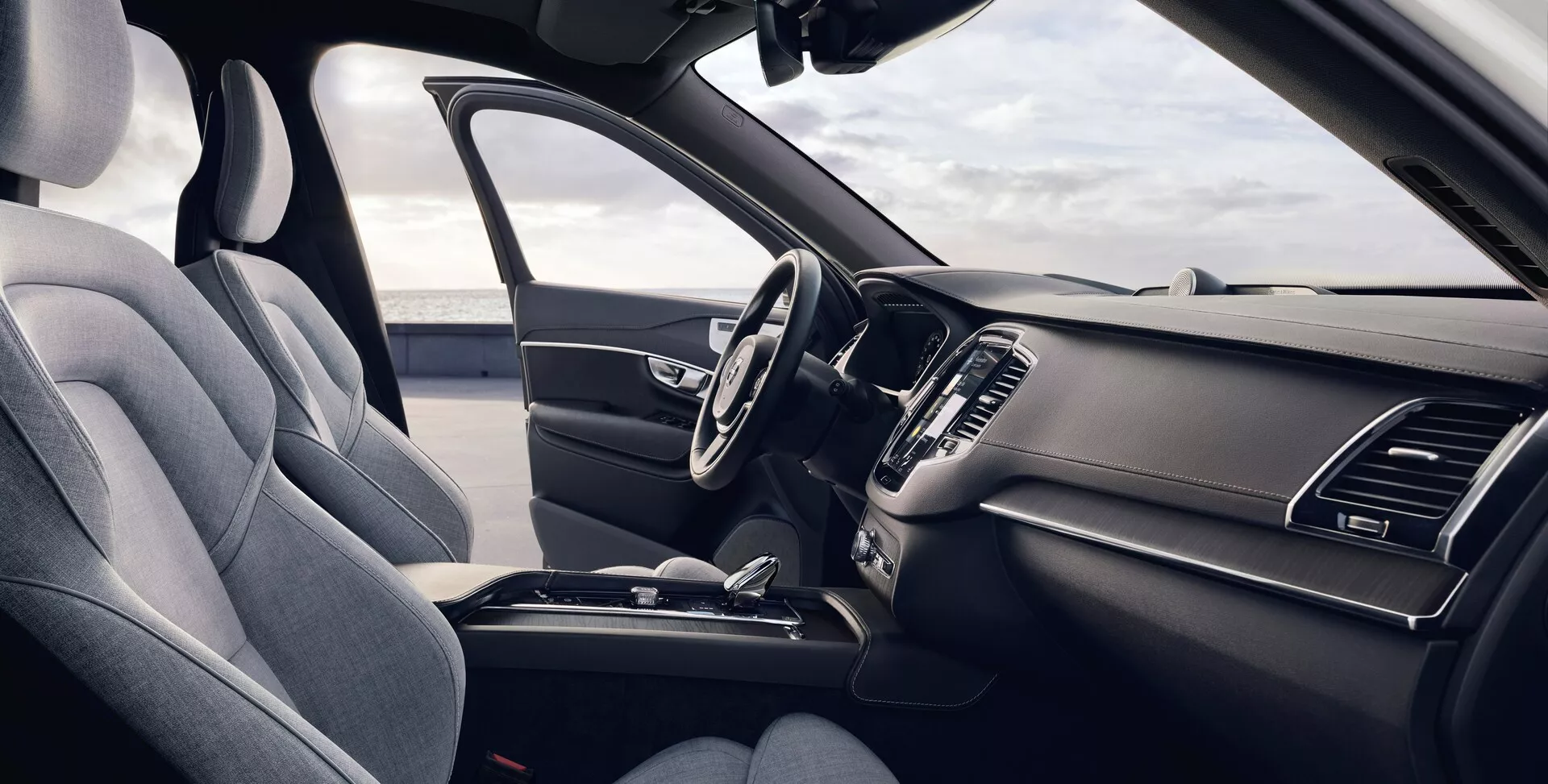 Vue intérieure d'une Volvo XC90 2015 (2e génération) depuis la perspective du passager avant, montrant le siège conducteur ergonomique avec une garniture en tissu foncé premium ou cuir synthétique, volant multifonction avec commandes intégrées, tableau de bord élégant avec système d'infodivertissement tactile éteint, bouches d'aération intégrées, commandes de climatisation minimalistes, console centrale avec rangement, porte-gobelets et levier de vitesse, le tout mis en valeur par un schéma de couleur sombre unifié avec la lumière naturelle illuminant le véhicule.