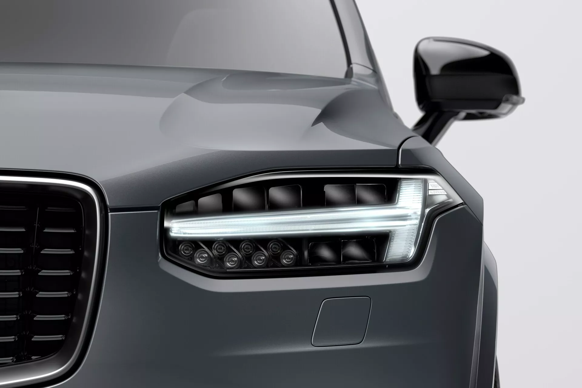 Vue de près du coin avant gauche d'une Volvo XC90 2015 (2e génération), montrant le design moderne des phares à LED avec des feux de jour angulaires, une vue partielle de la grande calandre à motif vertical, la peinture métallique gris foncé brillante et un capteur de stationnement sur le pare-chocs, prise à un angle bas de droite à gauche.