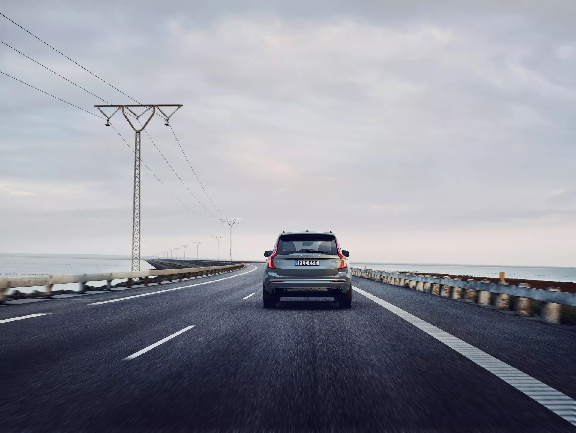 Vue arrière d'une Volvo XC90 de 2015 (2ème génération) circulant sur une route côtière à deux voies. La photo est prise sous un angle bas, avec une perspective centrée, capturant le hayon, la lunette arrière, les feux arrière, le pare-chocs arrière et les badge de marque et de modèle. Le véhicule est le principal sujet, avec un horizon océanique légèrement flou et des pylônes électriques en recul dans le lointain sous un ciel gris et couvert. La plaque d'immatriculation de la voiture est visible et l'éclairage suggère tôt le matin ou tard dans l'après-midi avec une lumière diffuse.