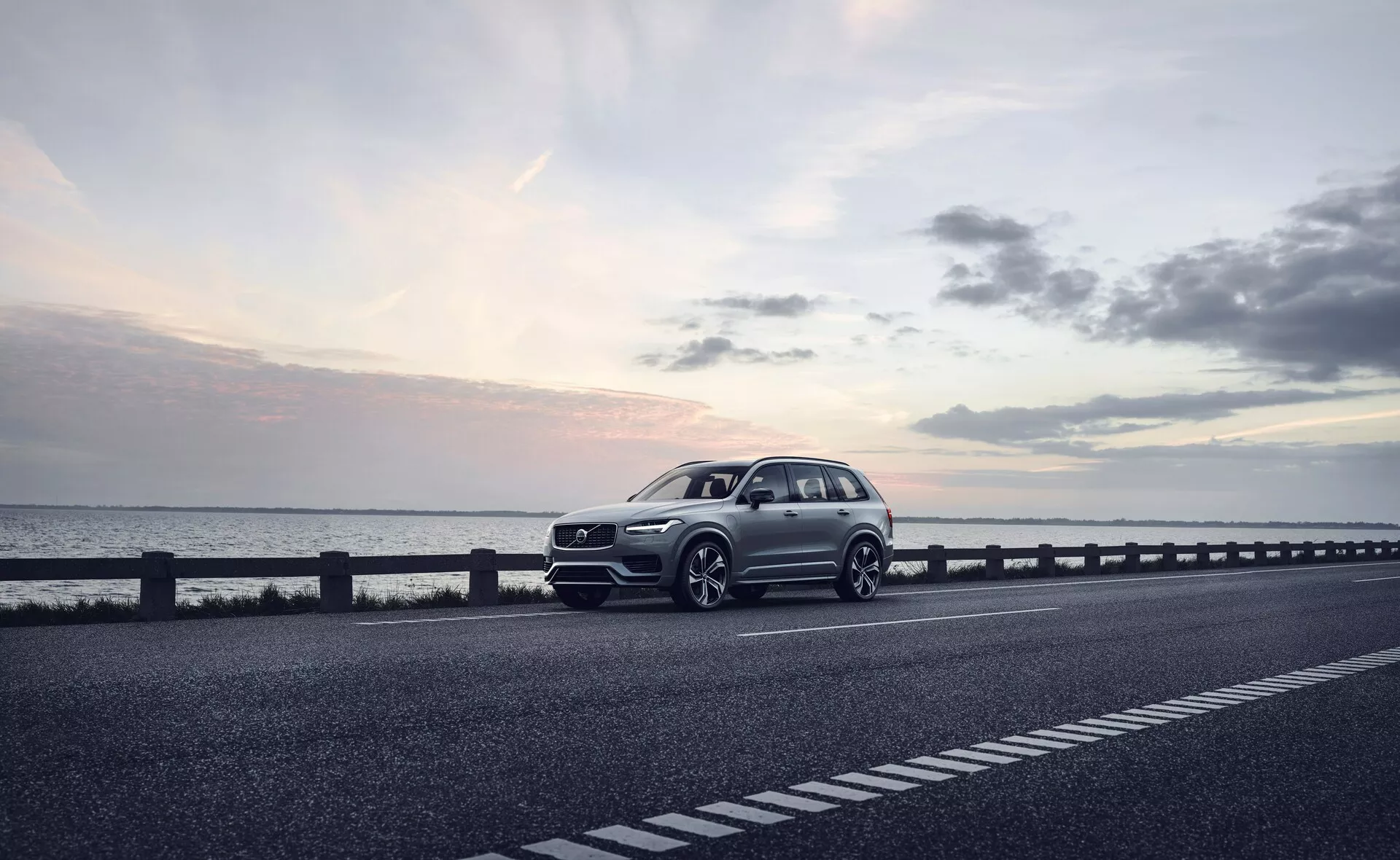 Un SUV moderne, spécifiquement le Volvo XC90 2015 (2ème génération), stationné sur le côté droit d'une route à deux voies. Le véhicule est capturé sous une vue de trois quarts avant, montrant la calandre avant, les phares, le capot et l'aile côté conducteur. L'angle révèle également une partie du profil de la voiture, y compris les roues, la porte du conducteur et une partie de l'arrière. Les roues sont légèrement tournées vers la droite, avec des éléments de design potentiellement sportifs comme un pare-chocs prononcé et des prises d'air plus grandes suggérant un pack dynamique. La photo, probablement prise au crépuscule avec une lumière douce et diffuse sous un ciel légèrement nuageux, renforce l'ambiance sereine. La carrosserie bien définie avec des lignes de caractère visibles indique un modèle haut de gamme. Les caractéristiques technologiques inférées de l'image incluent des phares à LED, des conceptions avancées de roues, une calandre avant sophistiquée avec des capteurs ou des caméras potentielles, et des touches aérodynamiques. Cette image de haute qualité pourrait être appropriée pour la publicité, mettant en avant la voiture comme un choix de style de vie luxueux.