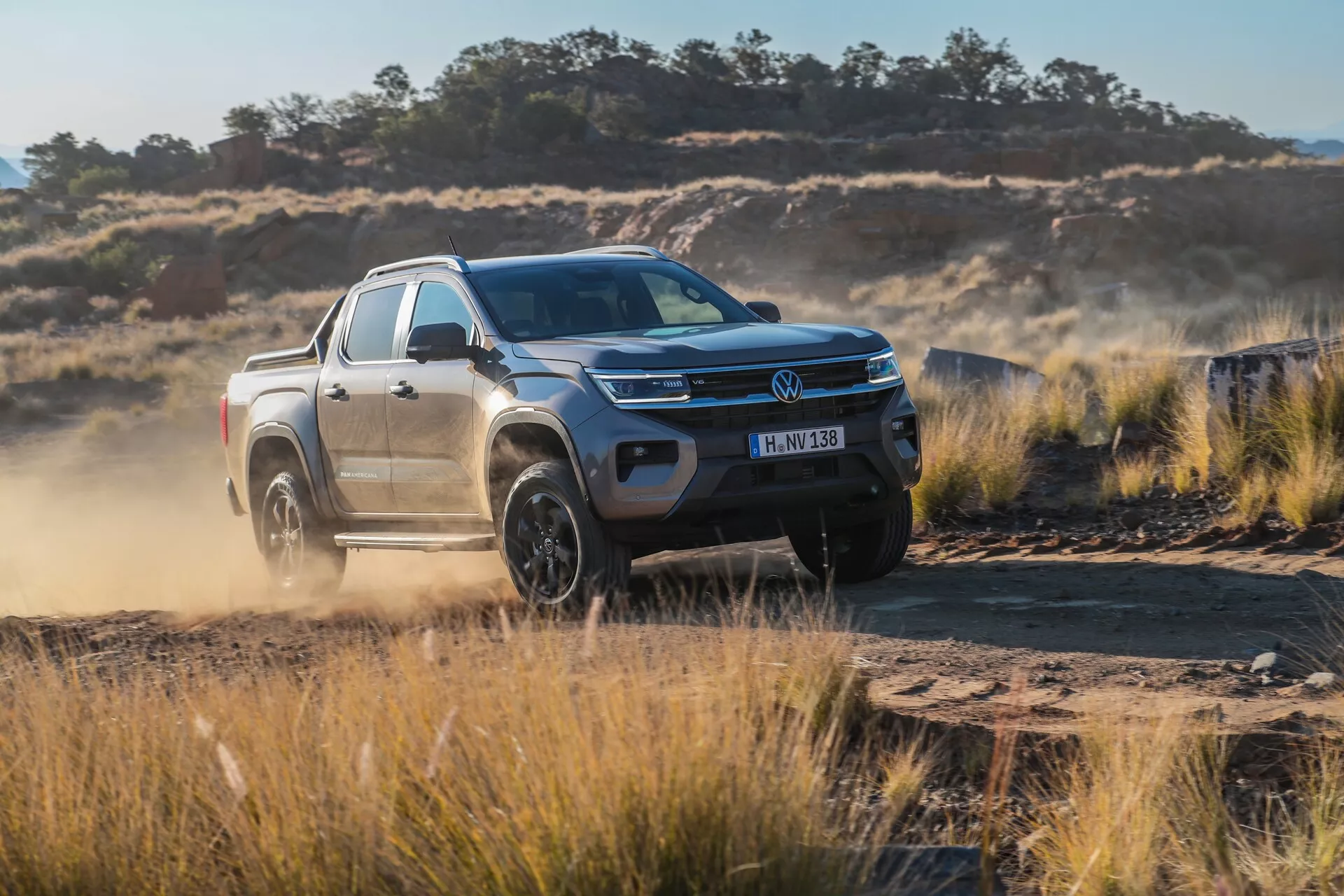 Vue de trois quarts avant du pickup VW Nutzfahrzeuge Amarok 2023 (2e génération), présentant son design dynamique sur une route en terre accidentée. L'image montre les phares à LED du camion, la calandre chromée avec l'emblème du fabricant, le capot sculpté, le pare-chocs avant robuste, les grandes jantes en alliage et les accessoires tels que les rétroviseurs latéraux avec clignotants intégrés, les barres de toit et l'arceau de sécurité. Le camion est représenté en mouvement, mettant en valeur ses capacités tout-terrain avec de la poussière soulevée derrière, suggérant des systèmes avancés de contrôle de traction et des capacités de conduite à quatre roues motrices.