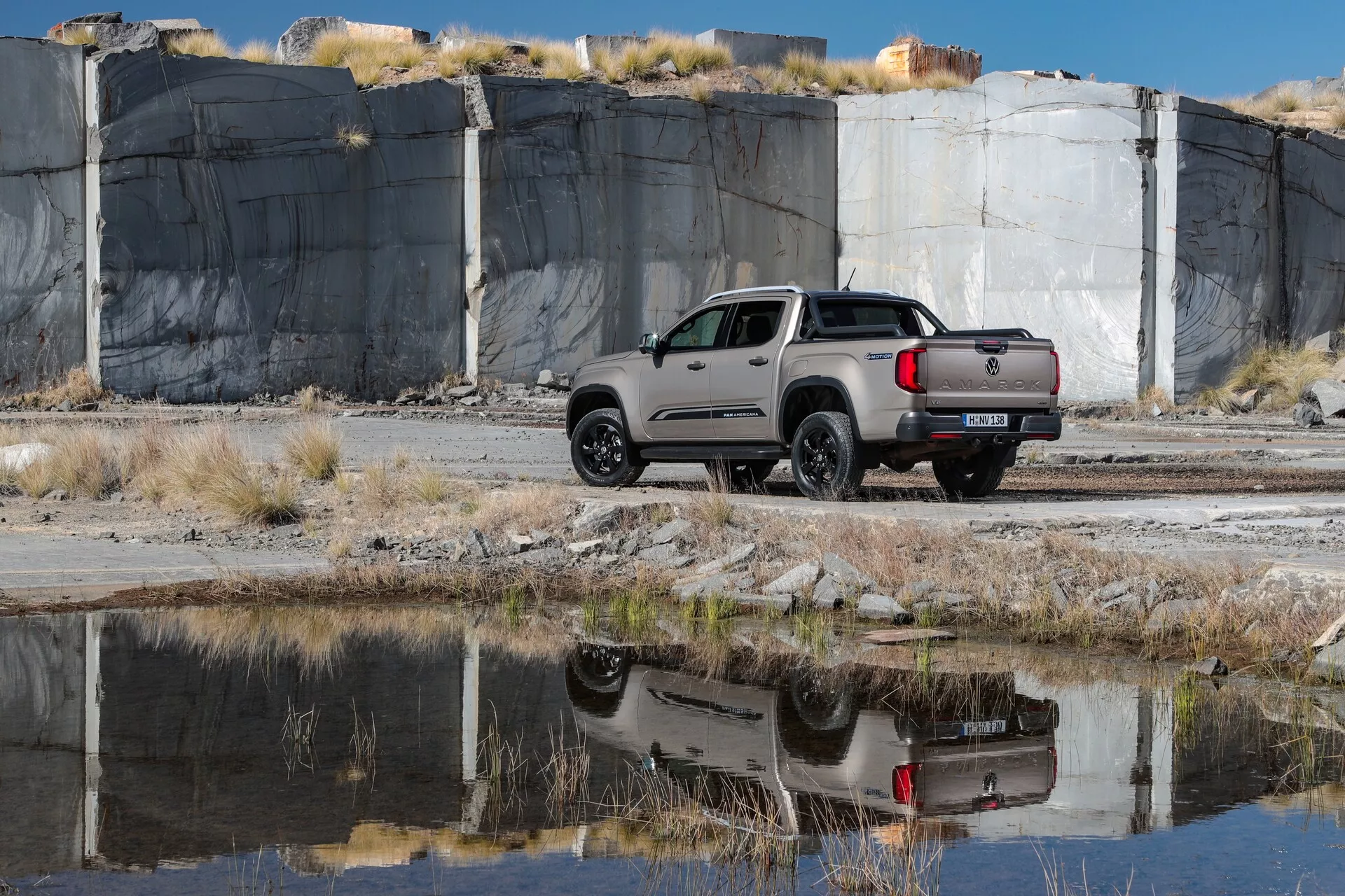 Voici une image du pick-up VW Nutzfahrzeuge Amarok 2023 de deuxième génération, photographié sous un léger angle latéral et arrière. Le côté droit du véhicule gris ou argenté ainsi que la partie arrière sont visibles, tandis que l'avant ne l'est pas. Le camion est positionné sur un sol en terre inégal avec des touffes d'herbe, probablement dans un cadre tout-terrain ou industriel, comme l'indique un grand mur de carrière de pierres en arrière-plan. La lumière naturelle illumine le camion, projetant une ombre douce vers la caméra. Les reflets du véhicule se voient sur la surface de l'eau au premier plan, capturant son profil latéral et arrière. Le camion présente un design moderne et élégant avec des feux arrière contemporains, des lignes de contour et des jantes en alliage. C'est une cabine double avec quatre portes et une benne courte, équipée d'un hayon moderne et de capteurs de pare-chocs arrière suggérant une technologie d'assistance au stationnement. Les rétroviseurs latéraux sont ajustés, indiquant des rétroviseurs latéraux réglables.