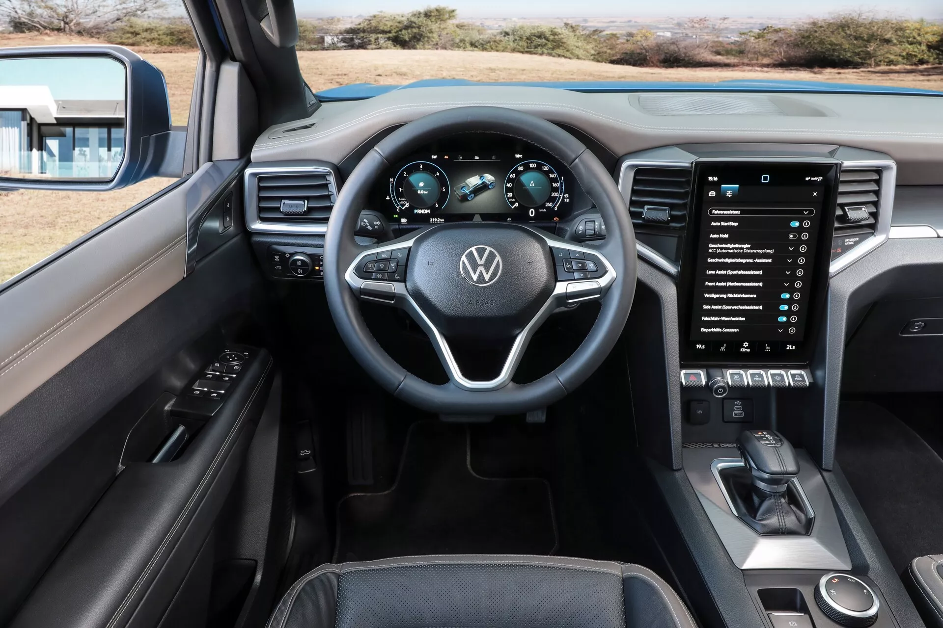 Vue intérieure depuis le siège du conducteur de la Volkswagen Amarok 2023 (2e génération) mettant l'accent sur le tableau de bord, le volant, le panneau d'instruments et le système d'infodivertissement. Le volant multifonctionnel avec le logo VW et les boutons de commande est centré, soutenu par un tableau de bord numérique affichant la vitesse et le régime moteur. Un grand écran tactile d'infodivertissement vertical domine la console centrale, entouré de boutons de contrôle du climat et d'un sélecteur de vitesses moderne, affichant un design de tableau de bord élégant et sophistiqué avec des matériaux de qualité.