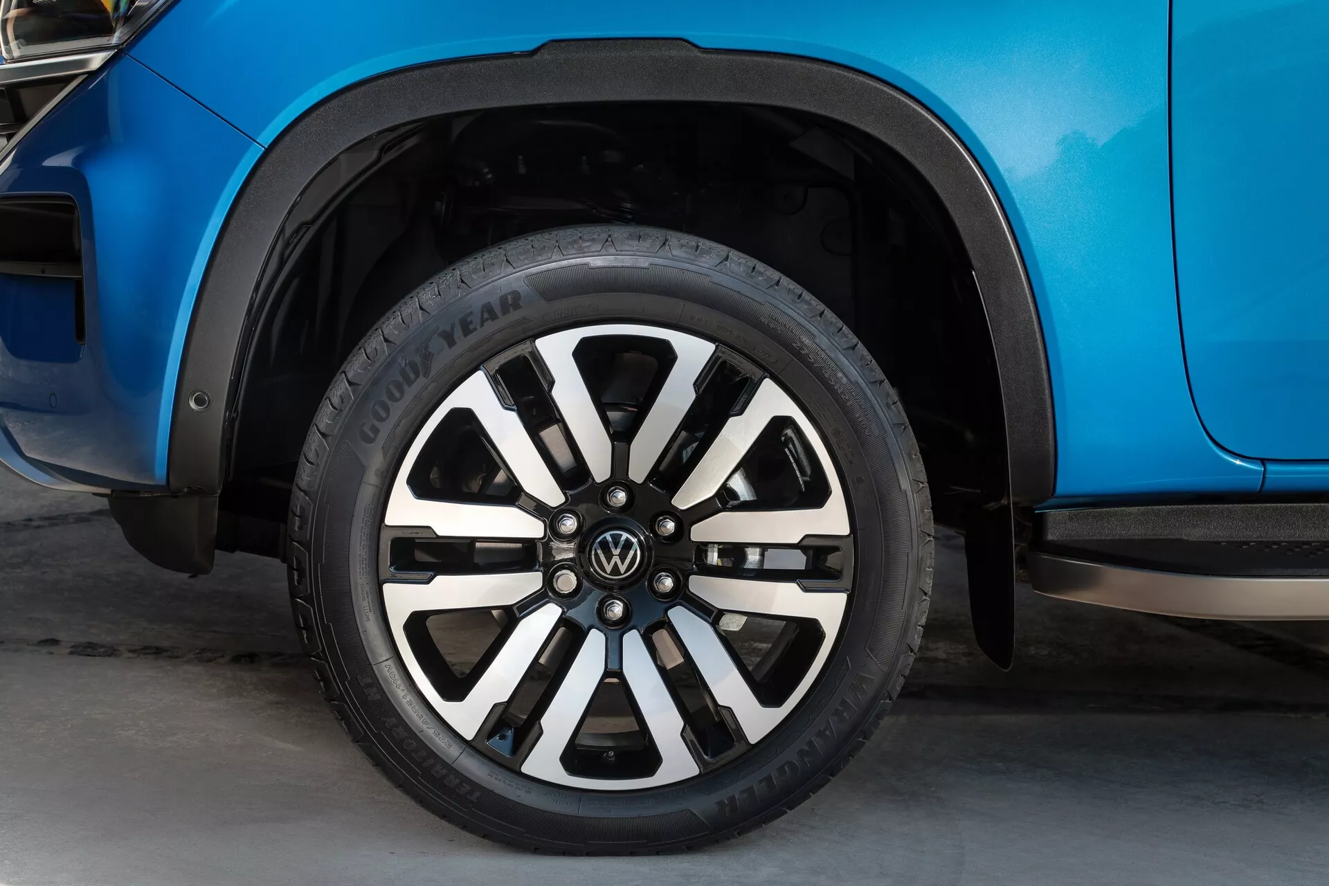 Vue rapprochée se concentrant sur la roue du VW Nutzfahrzeuge Amarok 2023 de deuxième génération. L'image montre le côté avant droit du véhicule, incluant une section de l'arche de la roue, le panneau de porte avant et les éclaboussures en plastique noir. La roue présente un design en alliage à rayons multiples avec des rayons alternant en noir et argenté avec un logo VW au centre. La bande de roulement du pneu Goodyear est visible et la finition de la peinture bleu métallique vibrant de la carrosserie du véhicule reflète la lumière. La photo est prise d'un angle bas, avec l'arrière-plan légèrement flou, mettant en évidence le pneu et la jante.