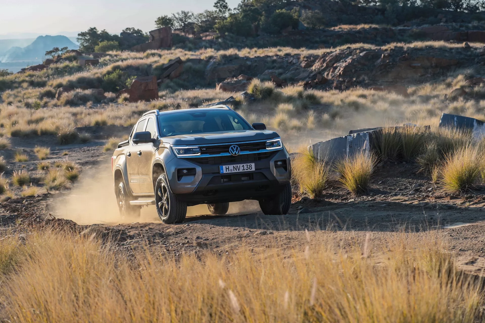 Volkswagen Amarok 2023 (2e génération) pickup capturé sous un angle de trois quarts avant dans un environnement tout-terrain. Le véhicule présente des éléments de design moderne tels que des feux diurnes à LED intégrés et un pare-chocs avant robuste. La photo met en évidence la garde au sol élevée de l'Amarok et le design de ses jantes en alliage, soulignant son aptitude pour les conditions difficiles. La poussière visible soulevée par les roues arrière montre ses capacités tout-terrain, le rendant idéal pour les aventuriers et ceux qui recherchent l'utilité et le style.