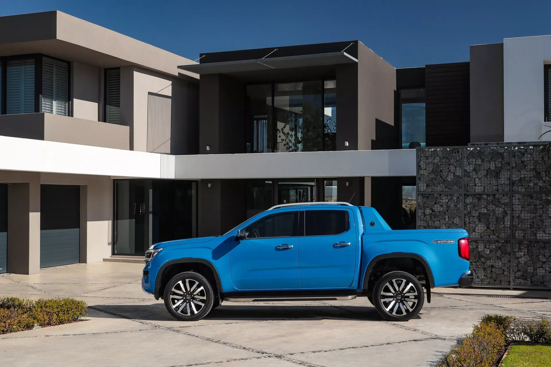 Image d'un VW Nutzfahrzeuge Amarok 2023 de deuxième génération, un pick-up moderne bleu avec peinture métallique, garé dans une allée résidentielle contre une toile de fond de maison de banlieue élégante. L'angle à trois quarts montre le profil du camion, y compris la forme de la cabine, du lit et des arches de roue, avec des indices sur la calandre avant et le design des phares. Les grandes roues en alliage suggèrent un mélange d'utilité et de luxe. La photographie est prise à une distance moyenne en lumière naturelle, soulignant l'apparence polie du véhicule et les caractéristiques de conception telles que les phares à LED, les technologies d'assistance au conducteur et une configuration de cabine double à quatre portes.